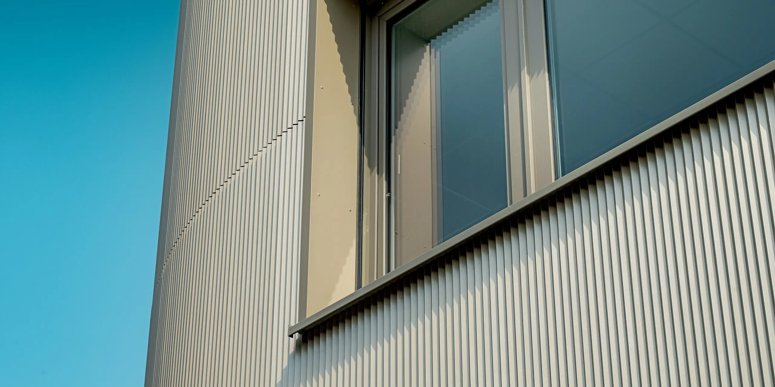 L'image révèle un détail de la façade du bâtiment Festo à Langenhagen, revêtue de profils extrudés triangle PREFA. La structure verticale des profilés crée une texture saisissante qui, associée à la pureté des encadrements de fenêtres, confère une esthétique moderne et dynamique. Le subtil jeu d'ombre et de lumière, créé par la façade en relief, renforce l'impact visuel du bâtiment et invite à en admirer les détails. La robustesse des matériaux PREFA garantit une surface durable et facile d'entretien, préservant ainsi l'esthétique architecturale de ce bâtiment pour les années à venir.