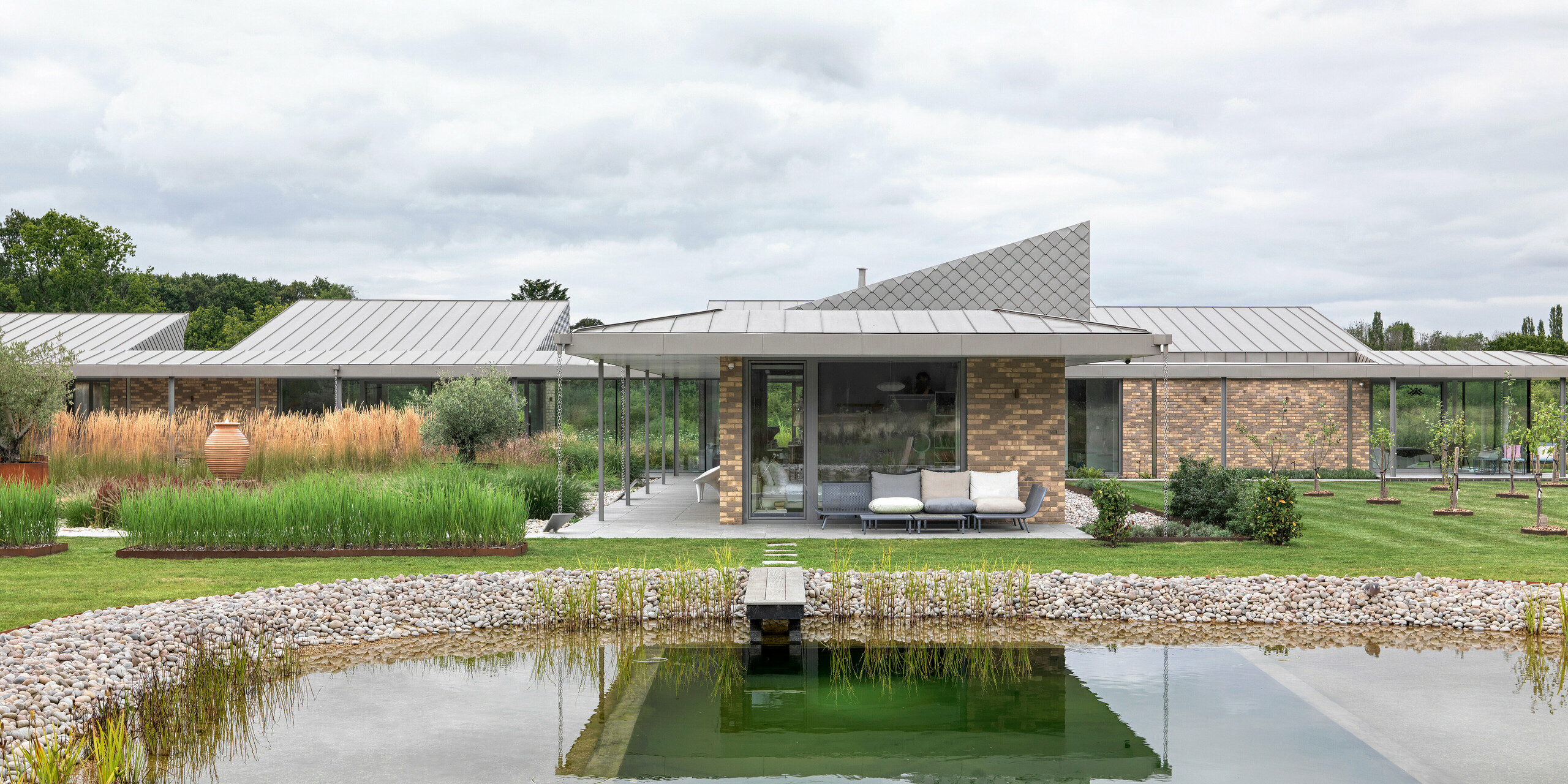 Vue d'ensemble symétrique d'une maison du West Sussex, avec sa toiture en aluminium PREFALZ couleur P.10 bronze et sa façade ornée de losanges PREFA 20 × 20. Au premier plan, une piscine naturelle paysagée avec une passerelle en bois, entourée d'un jardin paysager naturel. L'architecture épurée, avec ses larges baies vitrées et ses matériaux PREFA durables, crée un ensemble immobilier moderne, résistant aux intempéries et esthétique.
