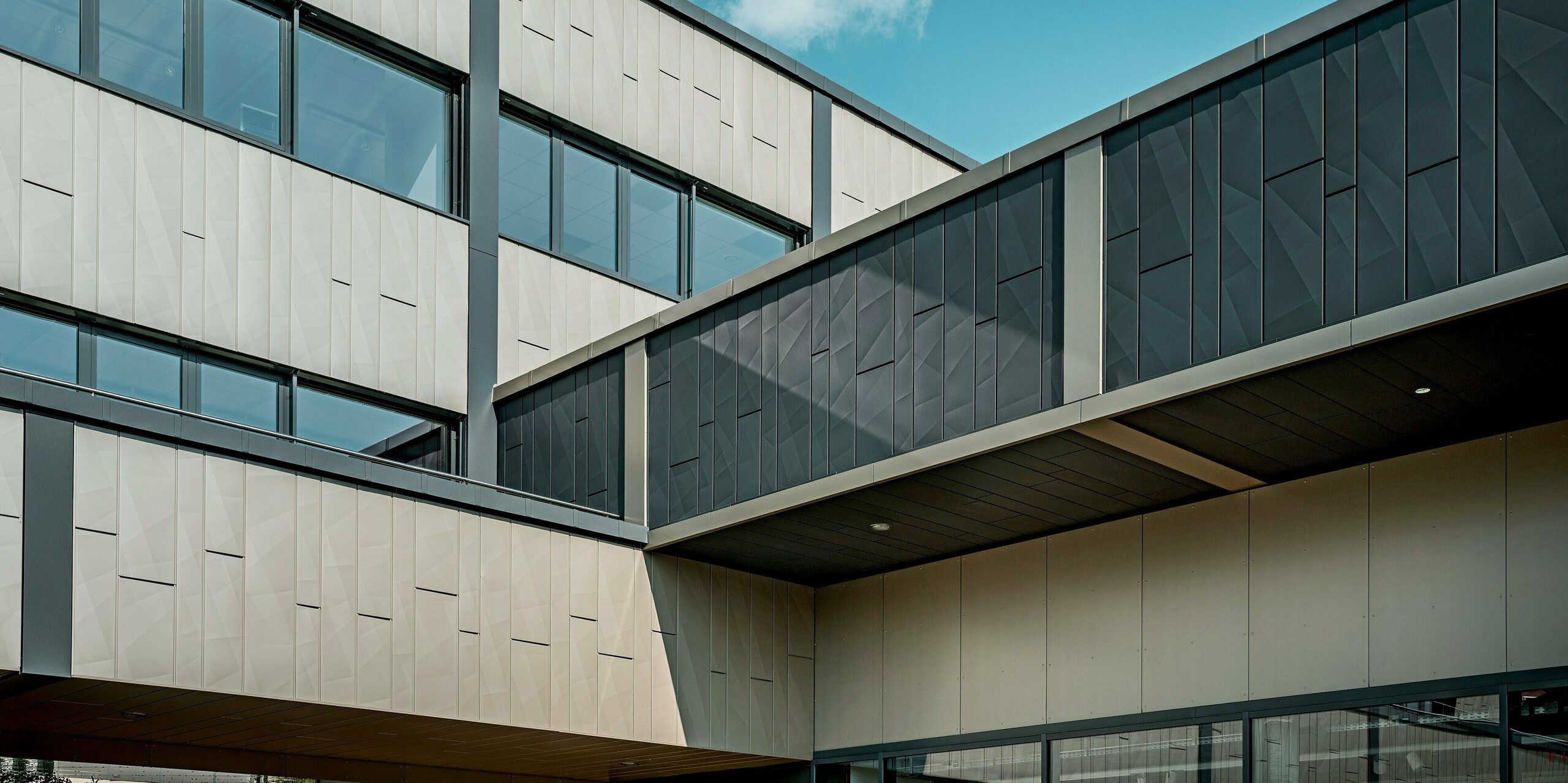 Die beeindruckende Aluminiumfassade des Einkaufszentrums in Rohrbach wurde mit PREFA Siding X-Fassadenpaneelen in den Farben Bronze und Anthrazit ausgestattet. Besonders auffällig ist der elegante Übergang zwischen den beiden Farben, der der Fassade eine dynamische und moderne Ausstrahlung verleiht. Die klare Struktur der Siding.X-Elemente sorgt für eine markante Gliederung der Wandflächen. Im Fokus stehen insbesondere die geometrischen Muster der Platten, die für eine besondere Tiefe sorgen.