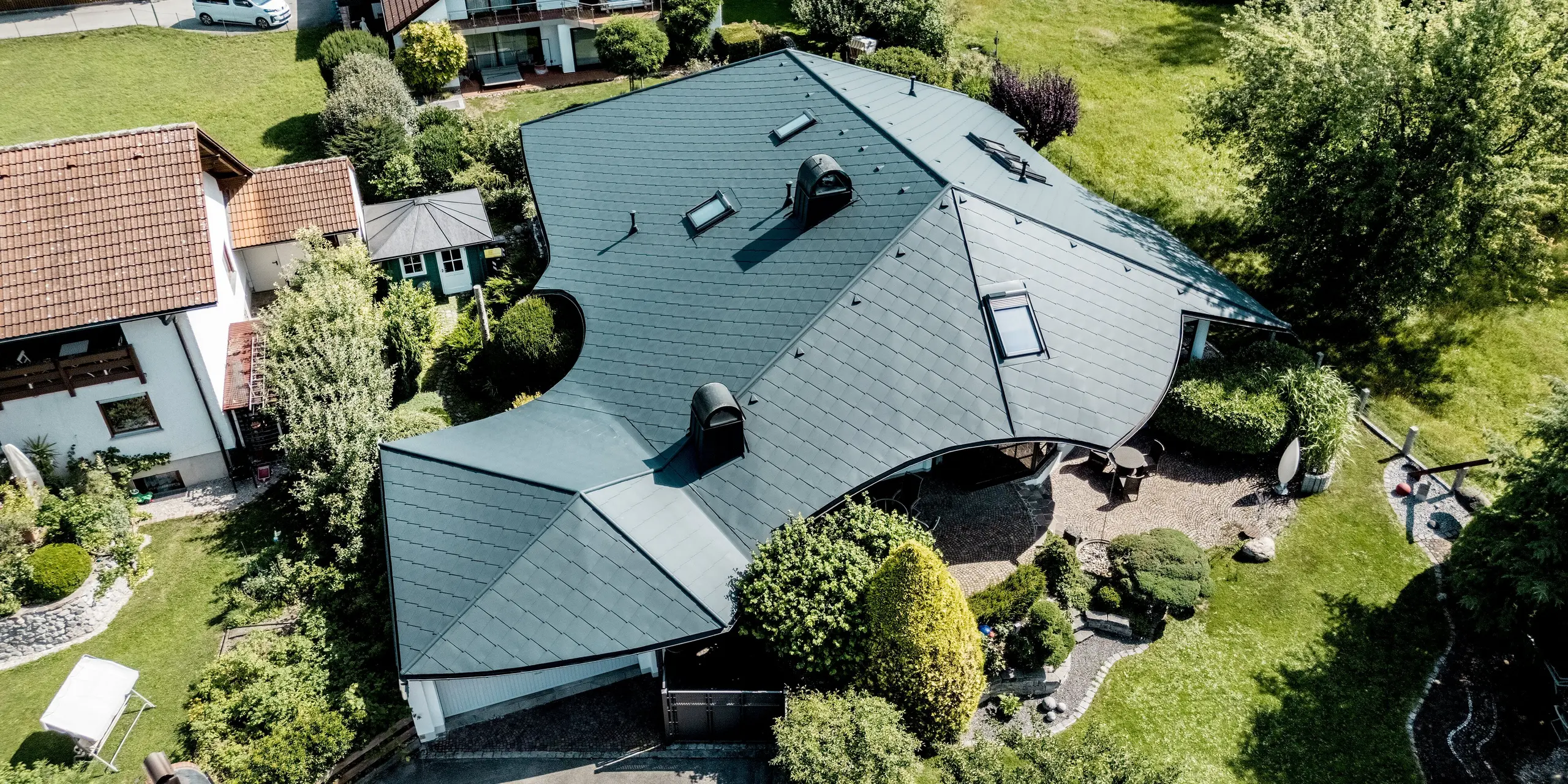 Drohnenaufnahme des sanierten Einfamilienhauses in Bobingen: Die außergewöhnlich geschwungene Dachlandschaft mit PREFA Dachraute 44 × 44 in P.10 Anthrazit zeigt zahlreiche Übergänge, Dachfenster und markante Aufbauten. Das leichte, robuste Aluminium passt sich der komplexen Geometrie präzise an und bietet langlebigen, witterungsbeständigen Schutz.