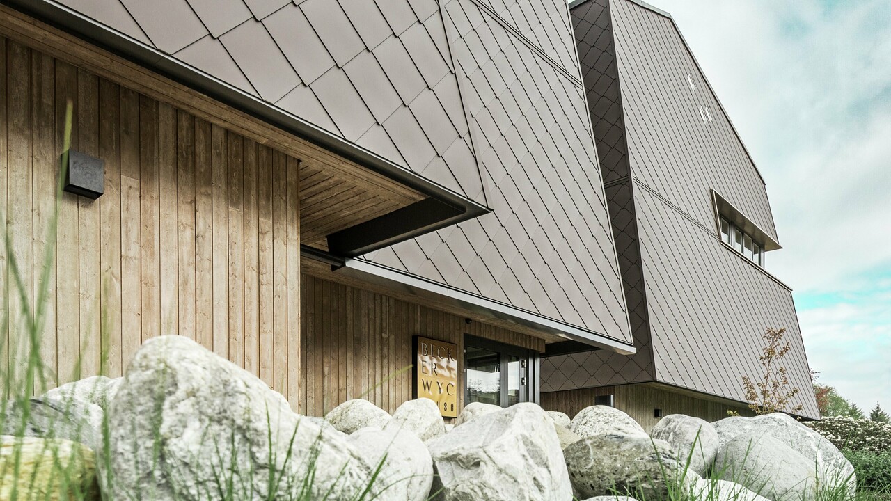 Vue latérale d'un hôtel, la façade du rez-de-chaussée est revêtue verticalement de bois plutôt sombre. Au-dessus se trouve un avant-toit, à partir duquel la façade est revêtue de plaques d'aluminium brun noisette.