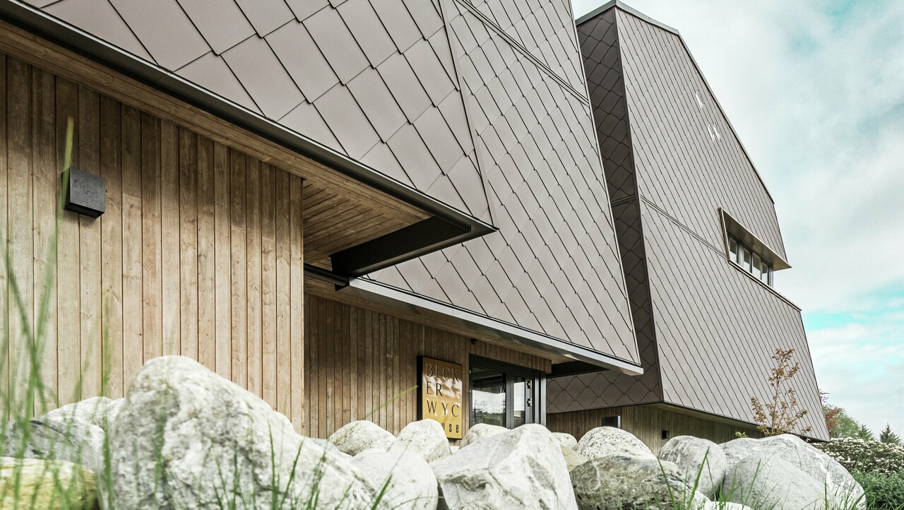 Seitliche Ansicht eines Hotels, die Fassade des Erdgeschosses ist mit eher dunklem Holz vertikal verkleidet. Darüber ist ein Fassadenvorsprung, ab hier ist die Fassade mit Aluminiumplatten in Nussbraun verkleidet.