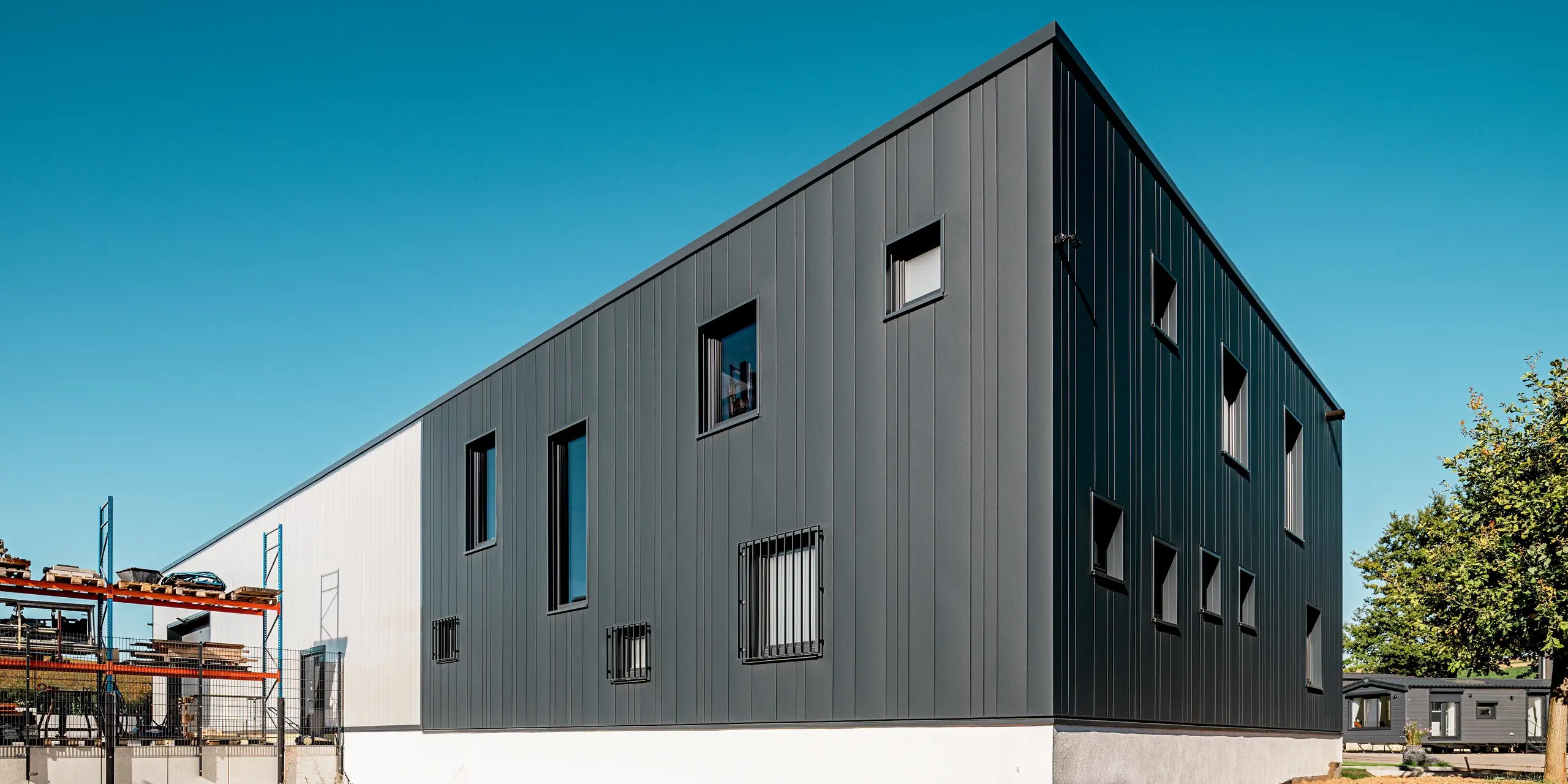 Cette image montre l'arrière du bâtiment Schädel GmbH, où les lignes verticales des Sidings PREFA sont particulièrement remarquables. La robuste surface P.10 anthracite confère à la structure compacte stabilité et élégance. Grâce à la variété des largeurs de panneaux, une façade dynamique est créée. La façade en aluminium PREFA offre une protection durable contre les intempéries et nécessite un entretien minimal.
