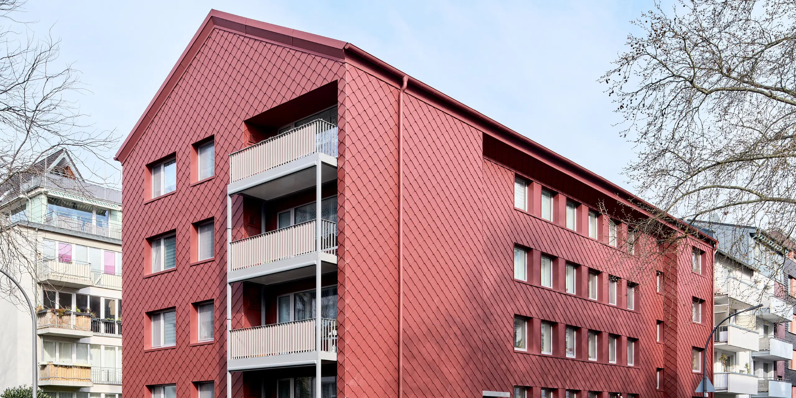 Architektonischer Blickfang in Köln-Zollstock: Das sanierte Mehrfamilienhaus „Energiesprong 2426“ beeindruckt mit einer durchgängigen Fassade aus PREFA Wandrauten 29 × 29 in P.10 Oxydrot. Das markante Rautenmuster umrahmt elegante Balkone und moderne Fensterreihen. Farblich abgestimmte PREFA Ablaufrohre und Saumrinnen runden das harmonische Gesamtbild ab. Die langlebige, witterungsbeständige Aluminiumfassade überzeugt durch Wartungsarmut.