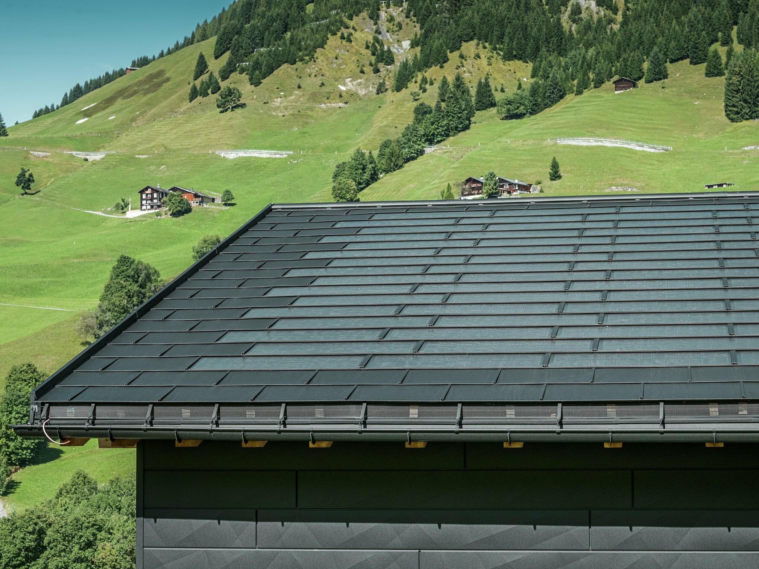 Gros plan sur l'avant-toit d'une maison familiale moderne dans les environs alpins d'Ascharina, en Suisse. Le toit a été recouvert de la tuile solaire innovante PREFA et du R.16 traditionnel, tous deux en P.10 noir. La gouttière suspendue PREFA et le tuyau de descente PREFA assurent un drainage fiable du toit. Les panneaux de façade distinctifs PREFA Siding.X sont visibles au bas de l'image. Les lignes claires des produits en aluminium soulignent le design moderne au milieu d'un paysage de montagne pittoresque.