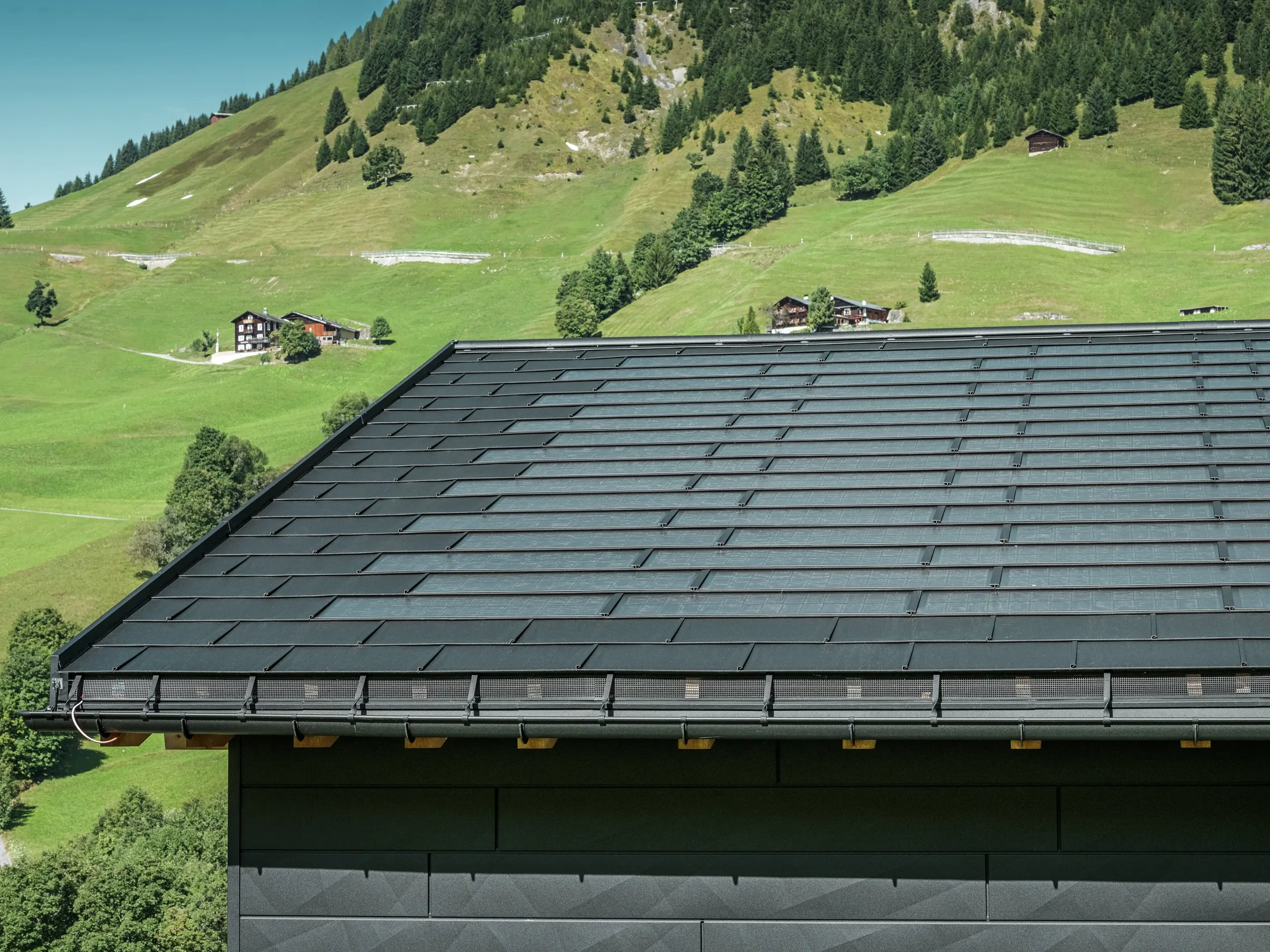 Nahaufnahme der Traufe eines modernen Einfamilienhauses in der alpinen Umgebung von Ascharina in der Schweiz. Das Dach wurde mit der innovativen PREFA Solardachplatte und der traditionellen Dachplatte R.16, beide in P.10 Schwarz, eingedeckt. Für die zuverlässige Dachentwässerung sorgen die PREFA Hängerinne und das PREFA Ablaufrohr. Am unteren Bildrand sind die unverwechselbaren PREFA Siding.X Fassadenpaneele zu sehen. Die klare Linienführung der Aluminiumprodukte unterstreicht das moderne Design inmitten einer malerischen Berglandschaft.