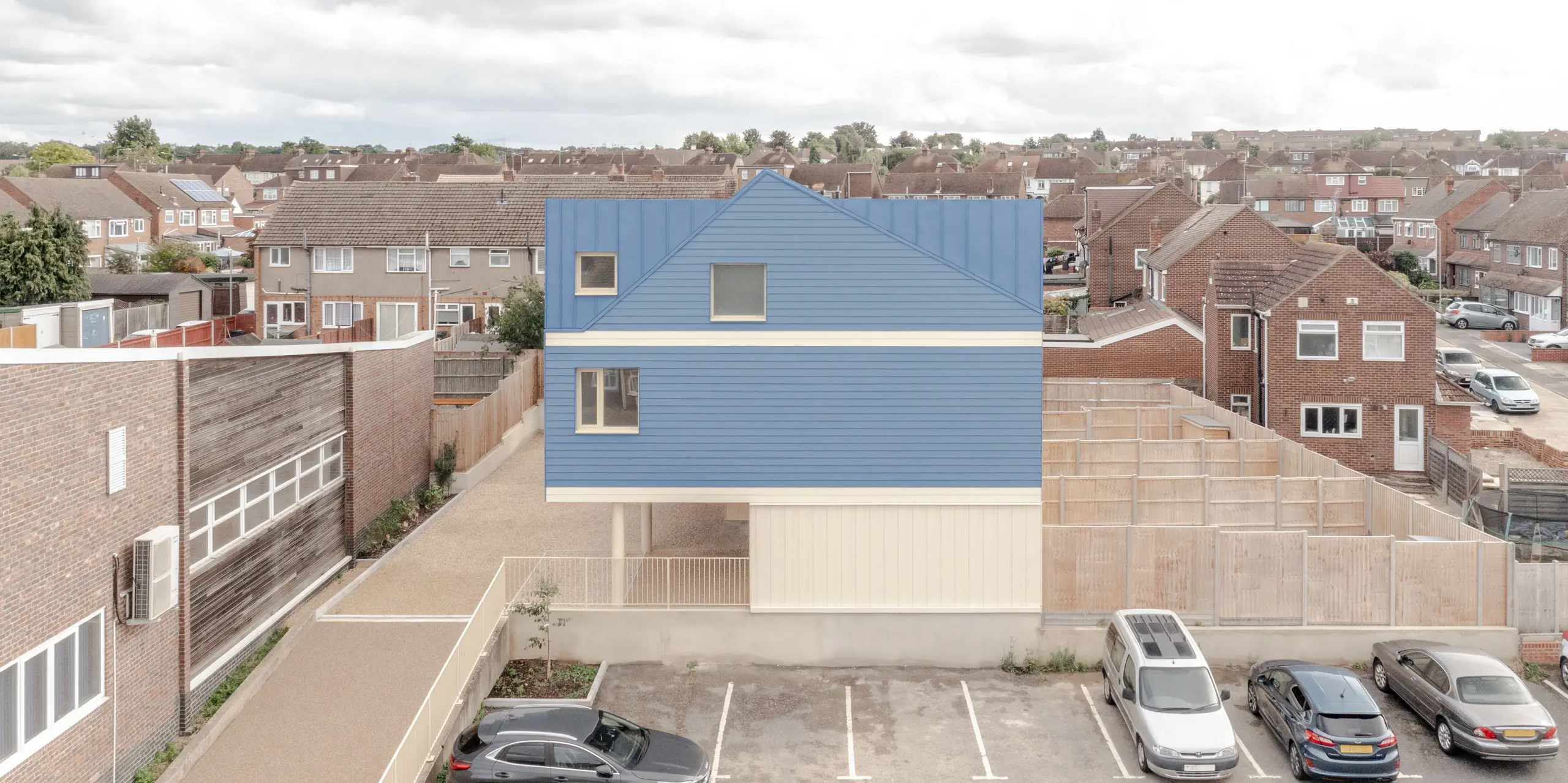 La photographie présente le complexe résidentiel Goldsmith-Mews en hauteur, mettant immédiatement en valeur la remarquable toiture et façade FALZONAL bleu pigeon, signées PREFA. La netteté des lignes et l'homogénéité de la structure confèrent au bâtiment une allure moderne et précise qui le distingue nettement des constructions environnantes. Le soubassement clair accentue cette impression de légèreté. L'aluminium PREFA, robuste et inaltérable, garantit une protection durable contre les intempéries et permet une conception architecturale à la fois résistante et expressive.