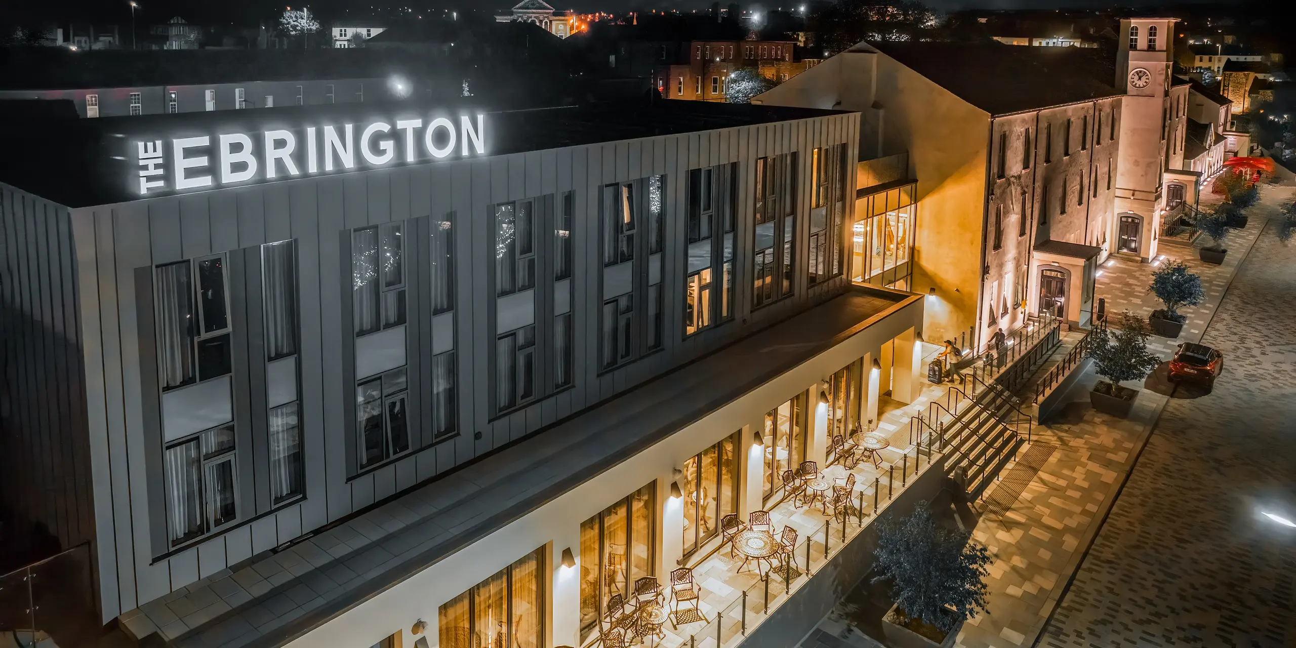 Das Ebrington Hotel bei Nacht besticht durch seine eindrucksvolle Beleuchtung in einer Fußgängerzone im Zentrum von Londonderry, Nordirland.  Das moderne Gästehaus wurde mit einer robusten PREFALZ Verkleidung aus Aluminium in den Farben P.10 Lichtgrau und P.10 Zinkgrau verkleidet. Diese hochwertige Blechfassade schützt das Gebäude vor Witterungseinflüssen. Zudem wirkt die Stehfalzdeckung als Highlight in der schlichten Architektur.