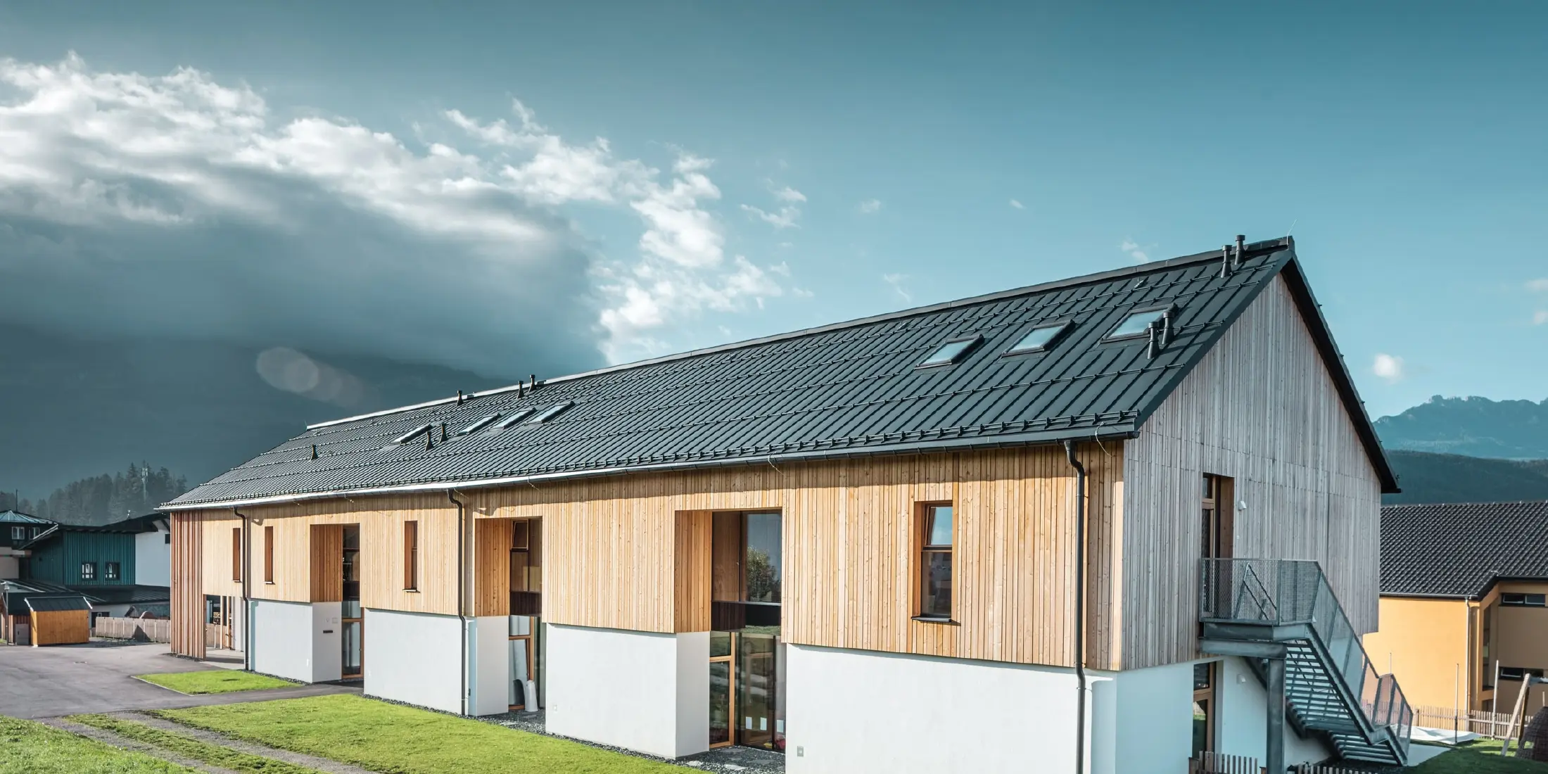 Modernes, langgestrecktes Gebäude mit Satteldach aus dunkelgrauem Aluminium und Fassade aus vertikaler Holzverschalung über weißem Sockel. Große Fensteröffnungen und eine Außentreppe aus Metall betonen die funktionale Architektur. Das Gebäude steht auf einer grünen Wiese mit umliegenden Häusern und Bergen im Hintergrund unter teils wolkigem Himmel.