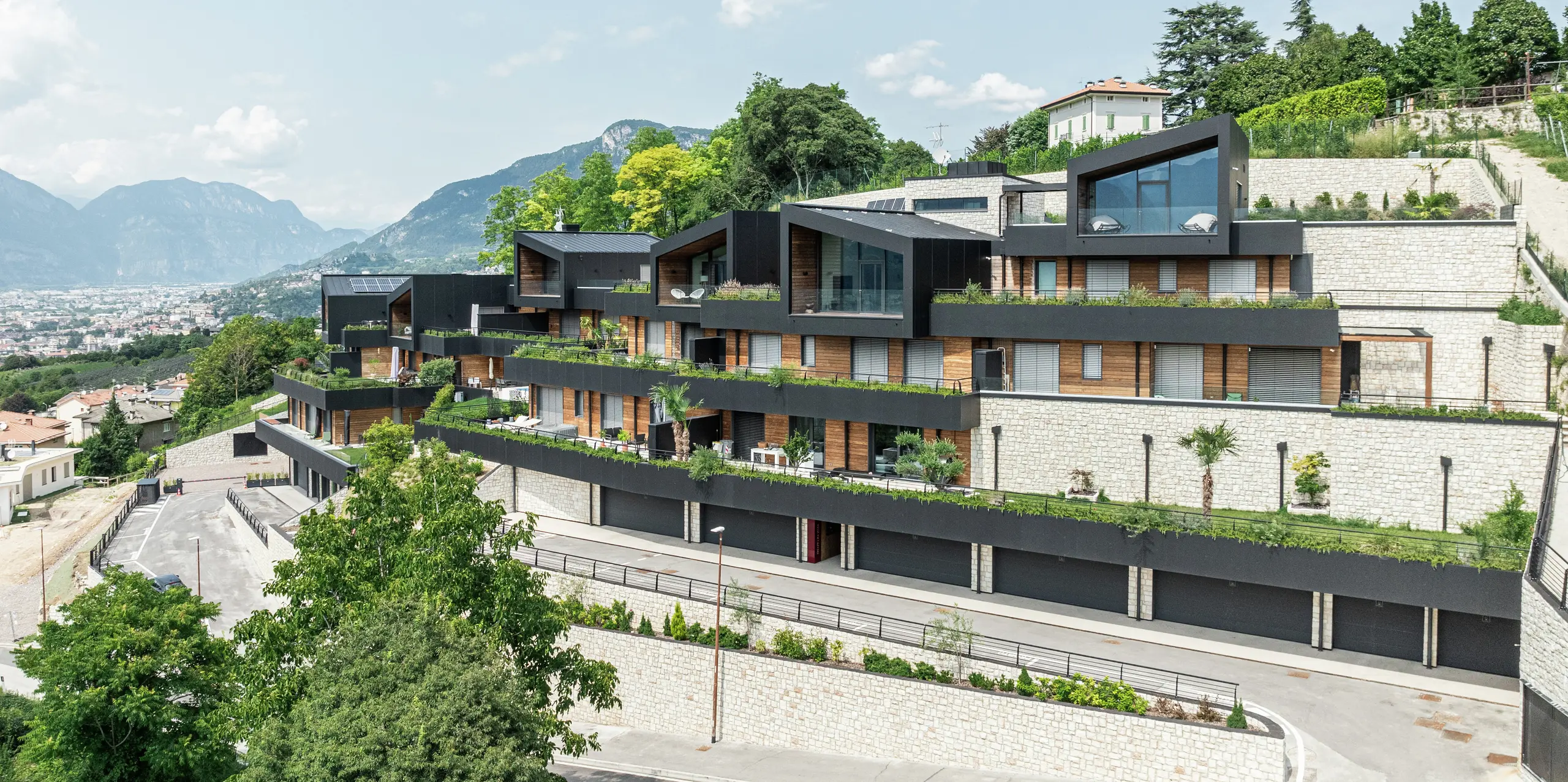 Le complexe résidentiel est parfaitement intégré dans la situation à flanc de colline au-dessus de Trente et offre une vue panoramique impressionnante sur la ville et le paysage montagneux environnant. La combinaison de la pierre naturelle, du bois et de la façade en aluminium PREFA de haute qualité en noir P.10 garantit une apparence moderne et élégante avec une résistance maximale aux intempéries. Le toit PREFALZ en P.10 Noir garantit durabilité et protection. Des terrasses spacieuses avec des plantes bien entretenues soulignent le concept de vie durable et créent une interaction harmonieuse entre l'architecture et la nature.