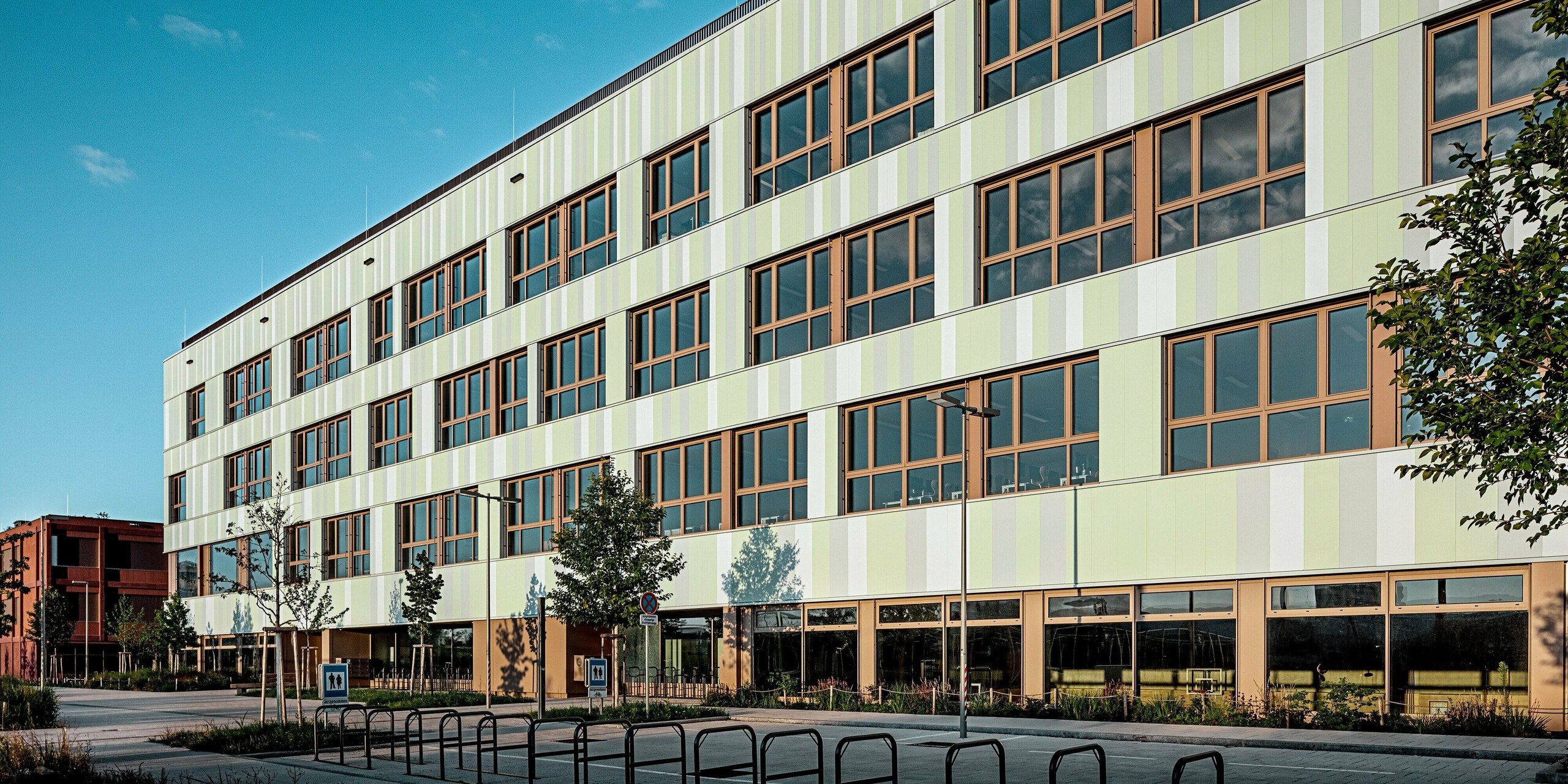 Die beeindruckende Fassade der AHS Reininghaus in Graz wurde mit PREFA Sidings in Sonderfarben ausgestattet. Die vertikalen Aluminiumprofile in sanften Grüntönen und Weiß verleihen der Schule eine frische und moderne Ausstrahlung. Besonders auffällig sind die Fensterrahmen in einem warmen Braunton, der einen schönen Kontrast zur helleren Fassadenfarbe bildet und das Gesamtbild harmonisch abrundet. Die präzise ausgerichteten Fenster und die gepflegten Grünflächen im Vordergrund sorgen für eine ansprechende und funktionale Gestaltung. Der Außenbereich ist mit Fahrradständern und Bäumen ausgestattet, was das nachhaltige und benutzerfreundliche Konzept des Gebäudes unterstreicht. 