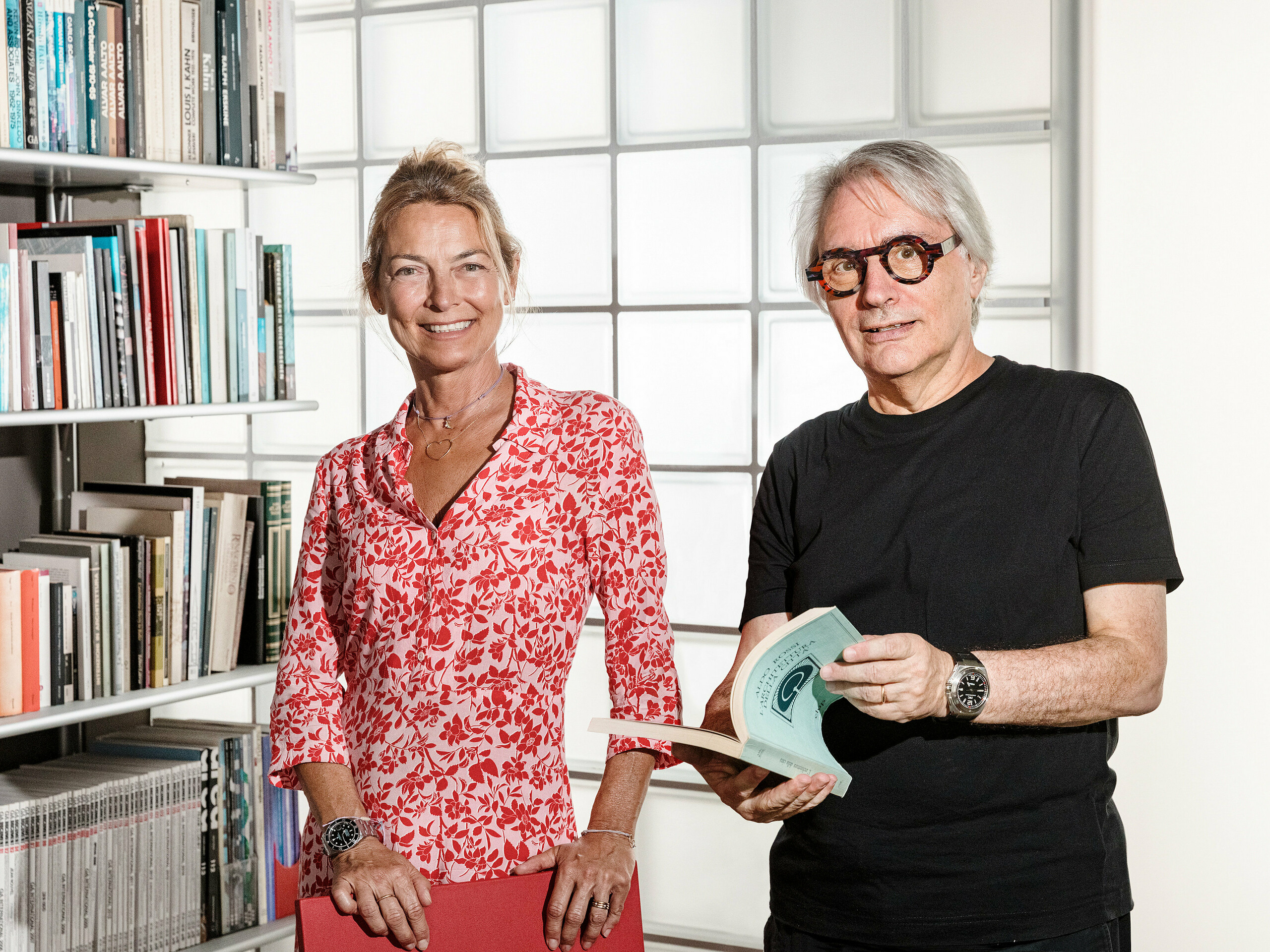 L'architecte Claudio Lucchin, originaire de Bolzano, et sa partenaire Daniela Varnier posent devant un mur moderne en briques de verre qui réfracte la lumière de manière fascinante. Claudio Lucchin, tout de noir vêtu, tient un livre dans ses mains. Daniela Varnier, vêtue d'un haut rouge vif, sourit aimablement. Ce décor élégant symbolise l'innovation et la transparence. La scène représente l'architecture d'avenir telle qu'elle a été réalisée dans la caserne « Tempesti » à Corvara, dans le Tyrol du Sud.