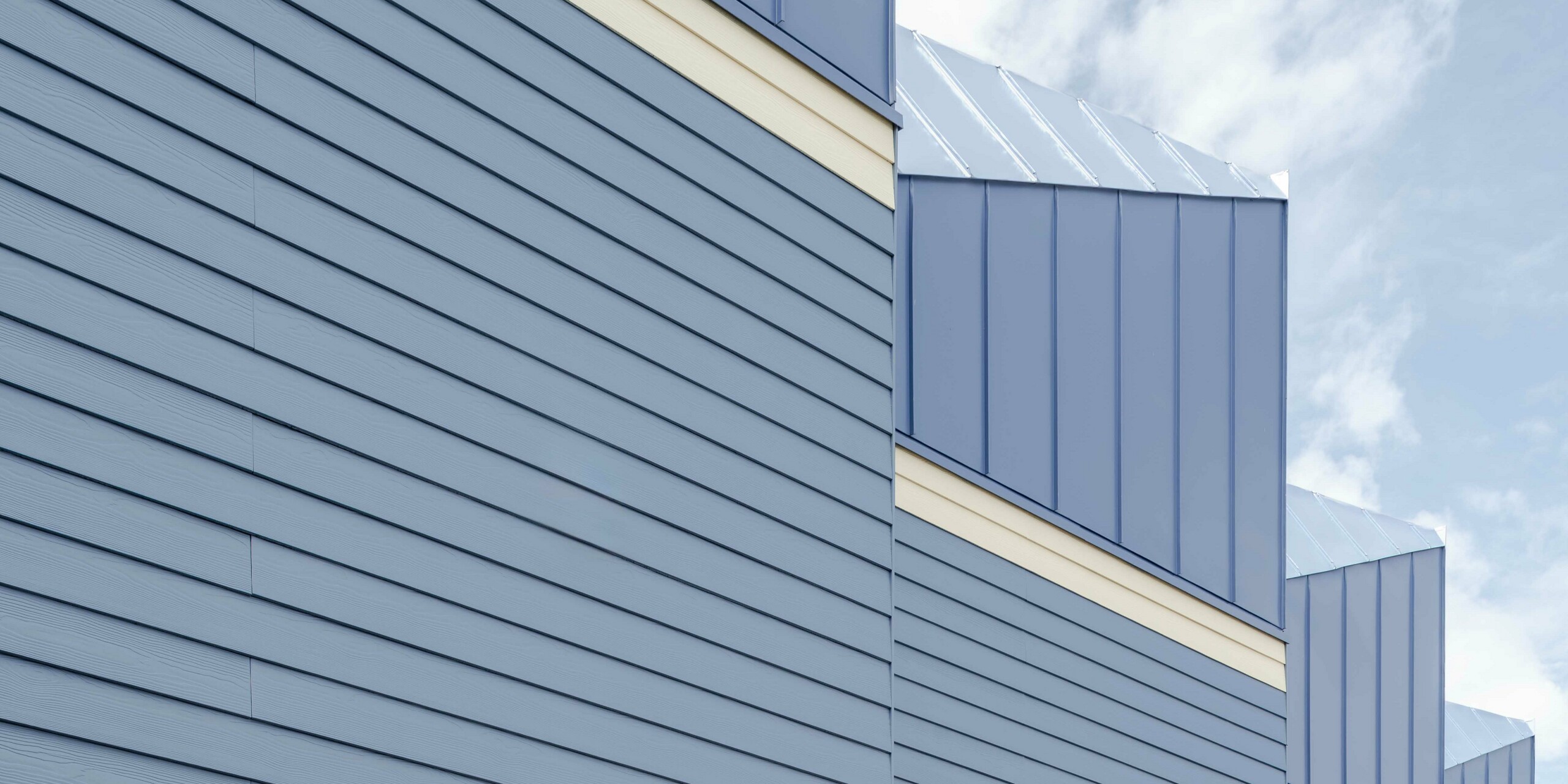 Die Aufnahme zeigt die markante Fassade der Goldsmith Mews Reihenhäuser mit ihrer auffälligen taubenblauen PREFA FALZONAL Aluminiumverkleidung. Die präzisen, vertikalen Linien der Aluminiumoberflächen erzeugen eine lebendige Struktur und betonen die moderne Architektur. Die hellen Untergeschosse verleihen dem Ensemble Leichtigkeit und verstärken den farblichen Kontrast. Das widerstandsfähige, farbstabile PREFA Aluminium schützt langfristig vor Witterung und ermöglicht eine klare, langlebige Gestaltung, die dem Projekt einen unverwechselbaren Charakter verleiht.