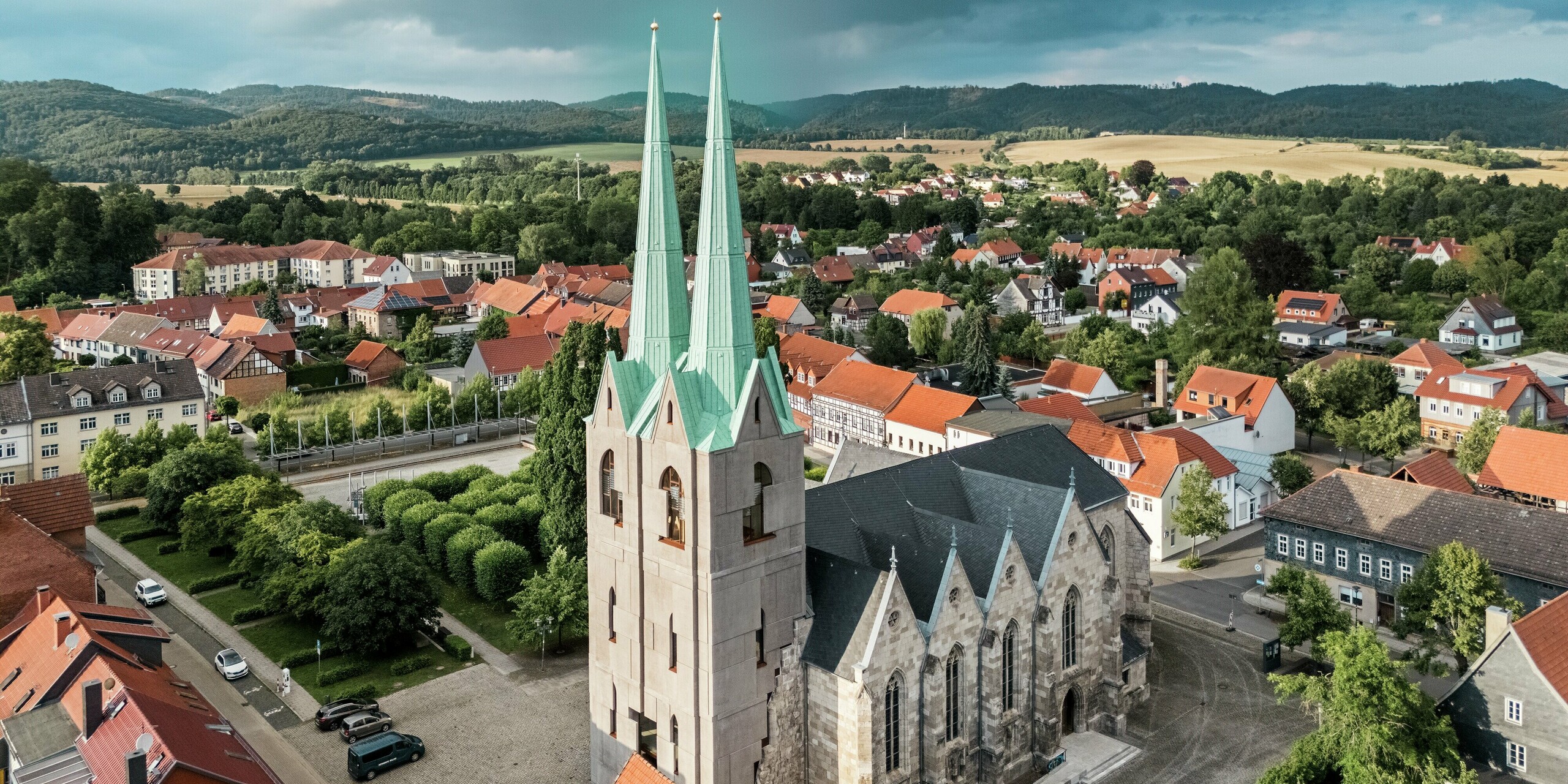 Luftaufnahme der sanierten Stadtkirche St. Johannis in Ellrich mit eindrucksvollen Doppeltürmen, eingedeckt mit PREFALZ in P.10 Patinagrün. Das elegante Aluminiumdach hebt sich klar von den roten Dächern der Umgebung ab und verleiht dem historischen Ensemble eine moderne Note. PREFALZ bietet dabei höchste Wetterbeständigkeit, Langlebigkeit und wartungsfreie Funktionalität.