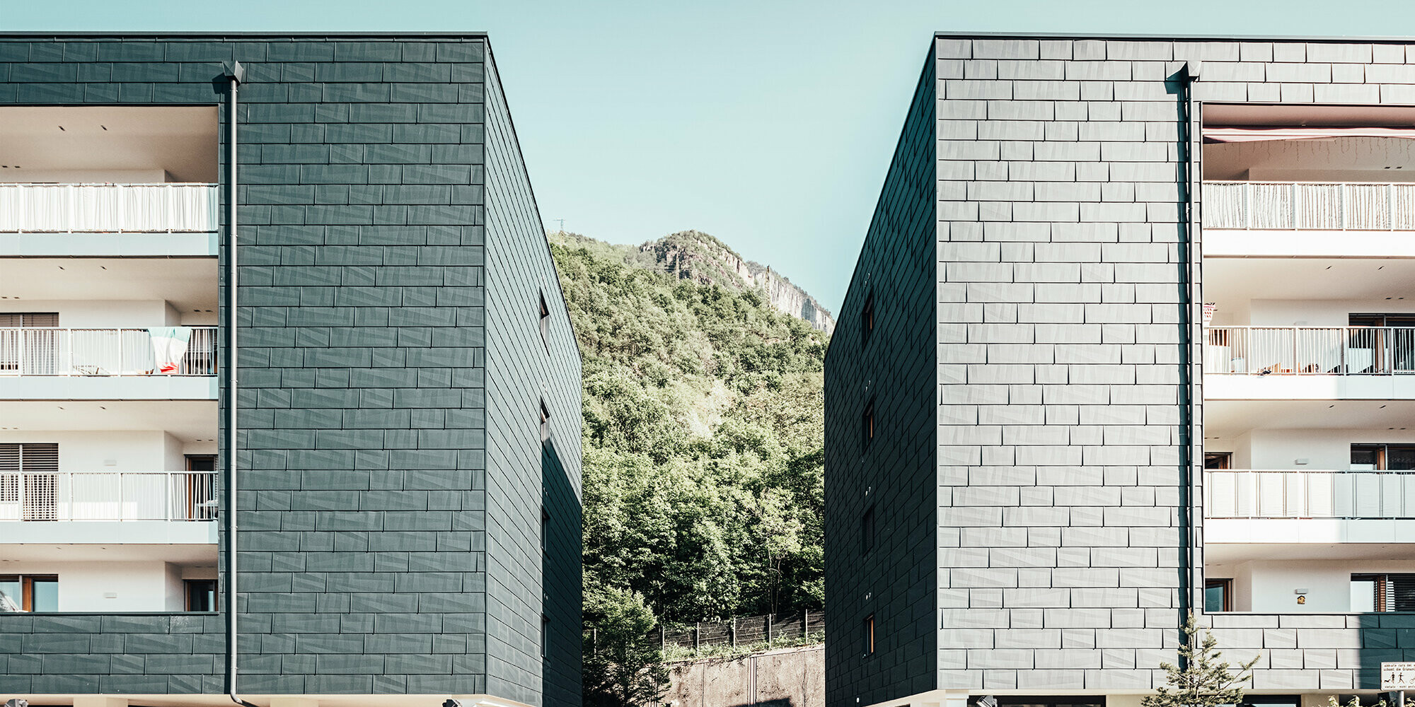 Prise de vue des deux côtés de l'immeuble aux façade à la teinte anthracite PREFA. Entre les deux, en arrière-plan, on aperçoit la montage et ses arbres.