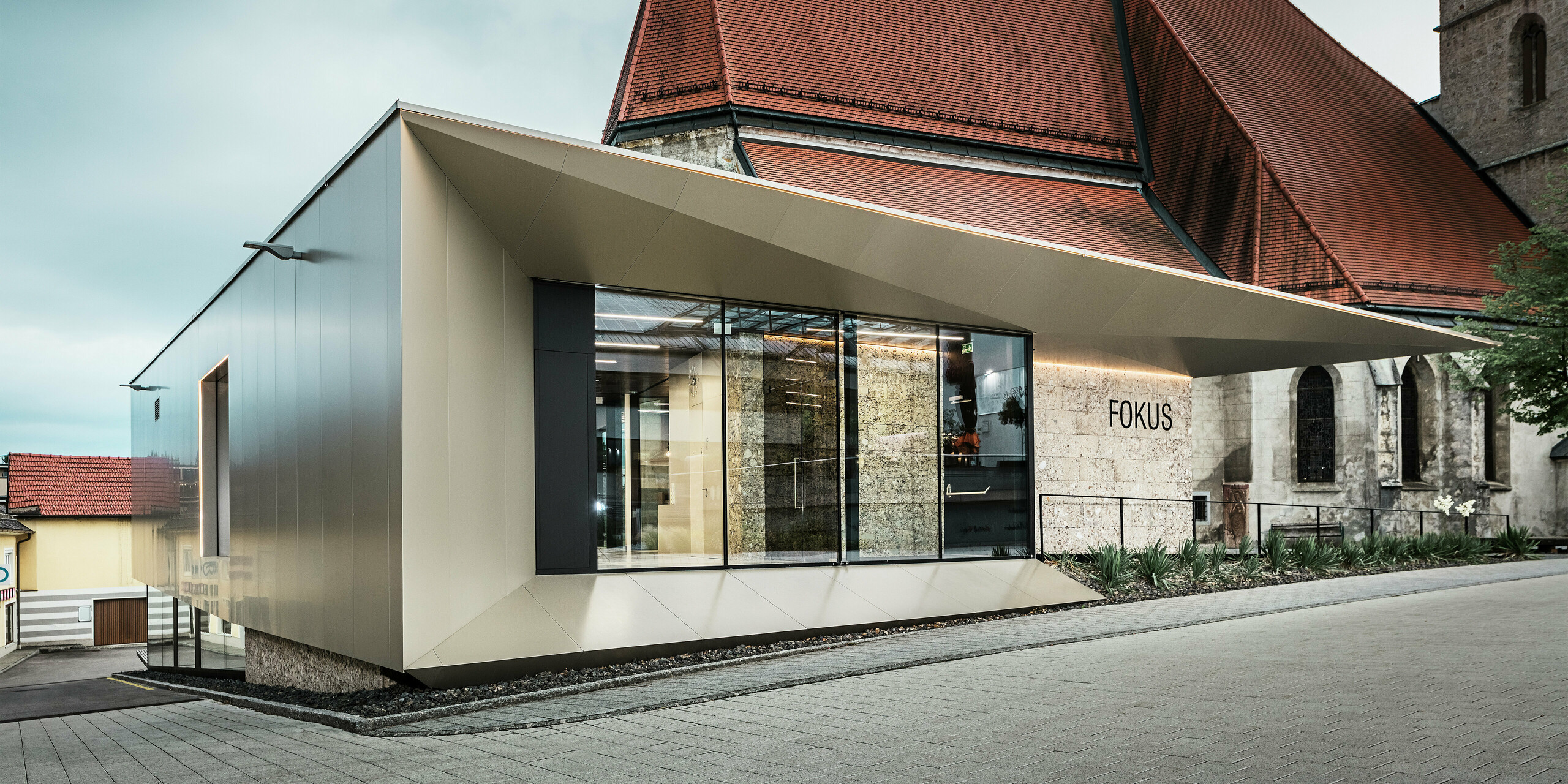 Im Vordergrund des Pfarrheims in Sierning glänzt die Aluminiumfassade durch ihre saubere Linienführung und hohe Formstabilität. Die Kombination aus Glasflächen und bronzefarbenen PREFA Aluminium Verbundplatten betont Offenheit, Beständigkeit und gestalterische Präzision bei gleichzeitiger Integration ins historische Umfeld.