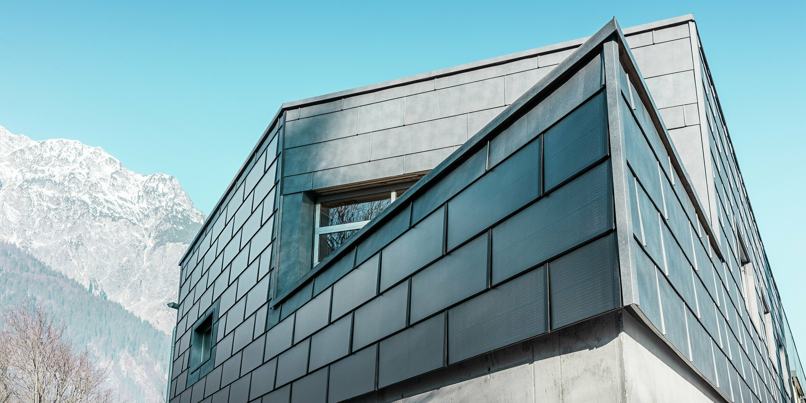La façade du bâtiment de l'entreprise de couverture RÜMA, dans le Vorarlberg, est ornée d'élégantes tuiles solaires PREFA et de panneaux de façade PREFA FX.12 associés à du béton apparent, sur fond de ciel bleu azur et de montagnes. L'angle prononcé du bâtiment souligne son architecture remarquable.