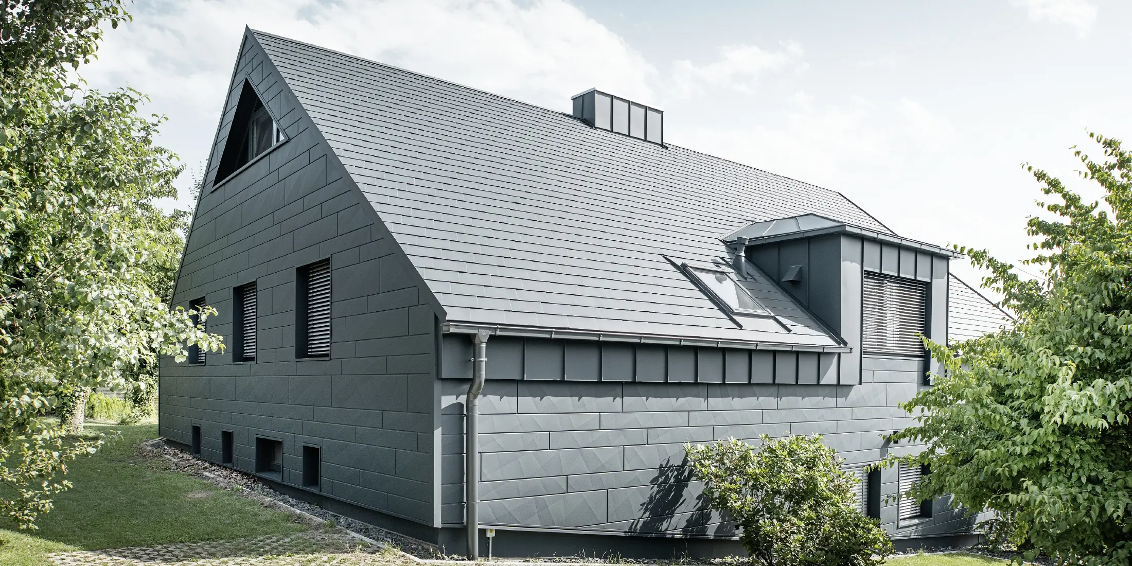 Grote eengezinswoning met zadeldak en veel ramen. Het hele huis is bekleed met een aluminium omhulsel in antraciet, op het dak zijn PREFA aluminium dakspanen gebruikt en op de gevel Siding.X in antraciet.