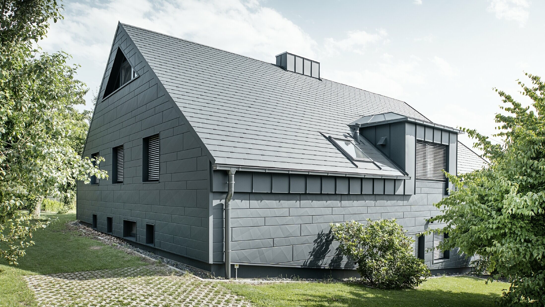 Grote eengezinswoning met zadeldak en veel ramen. Het hele huis is bekleed met een aluminium omhulsel in antraciet, op het dak zijn PREFA aluminium dakspanen gebruikt en op de gevel Siding.X in antraciet.
