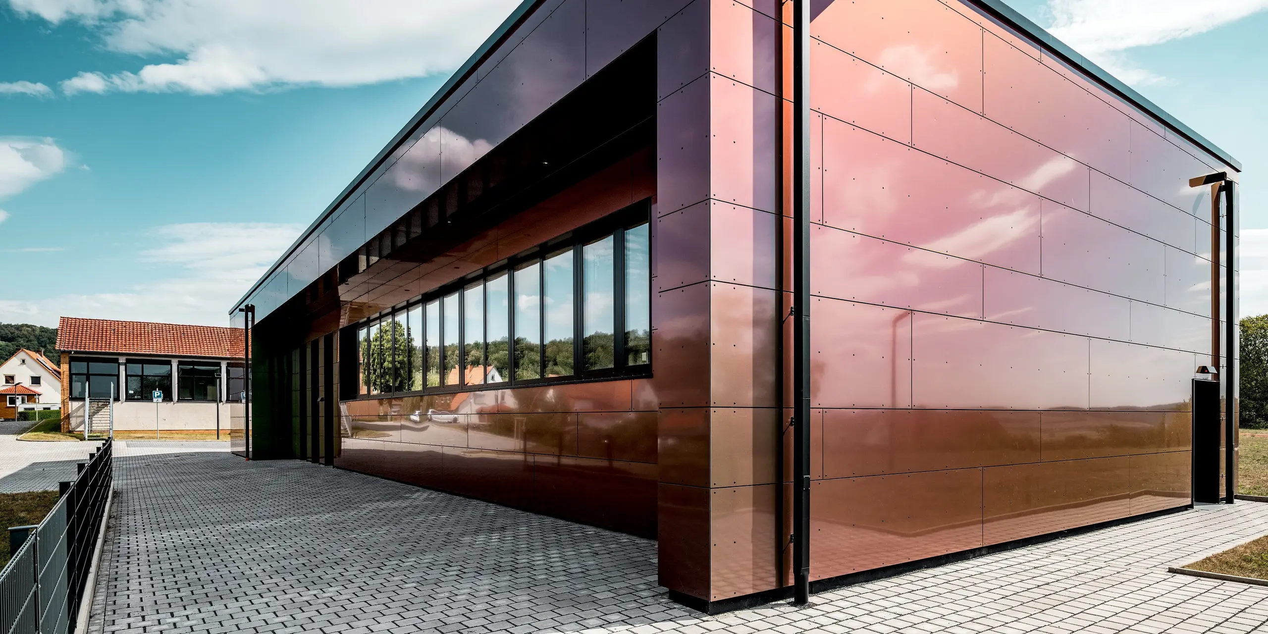 Beeindruckende Perspektive auf die Johannwolfschule Angerstein: Die glänzende PREFA Fassade aus großformatigen Aluminium Verbundplatten in der Sonderfarbe Spring Lake schimmert im Sonnenlicht in faszinierenden Rosa-, Bronze- und Violetttönen. Die spiegelnden Oberflächen, langen Fensterfronten und klaren Gebäudekanten prägen die moderne Architektur. Das dunkle PREFA Quadratrohr fügt sich harmonisch in die Fassadengestaltung ein.