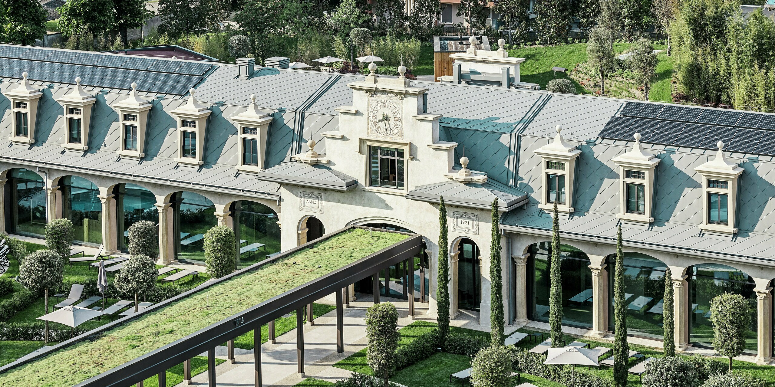 Cet élégant bâtiment Art nouveau restauré, situé dans le quartier milanais de San Siro, impressionne par sa toiture remarquable en losanges PREFA 44 x 44 et à joint debout PREFALZ, sous l'imposante installation photovoltaïque. Ces deux produits en aluminium sont peints en P.10 gris souris. Au premier plan, une arcade paysagère est entourée d'un jardin soigné, agrémenté de chaises longues, de parasols ombragés, de cyprès et d'allées sinueuses offrant un confort optimal.