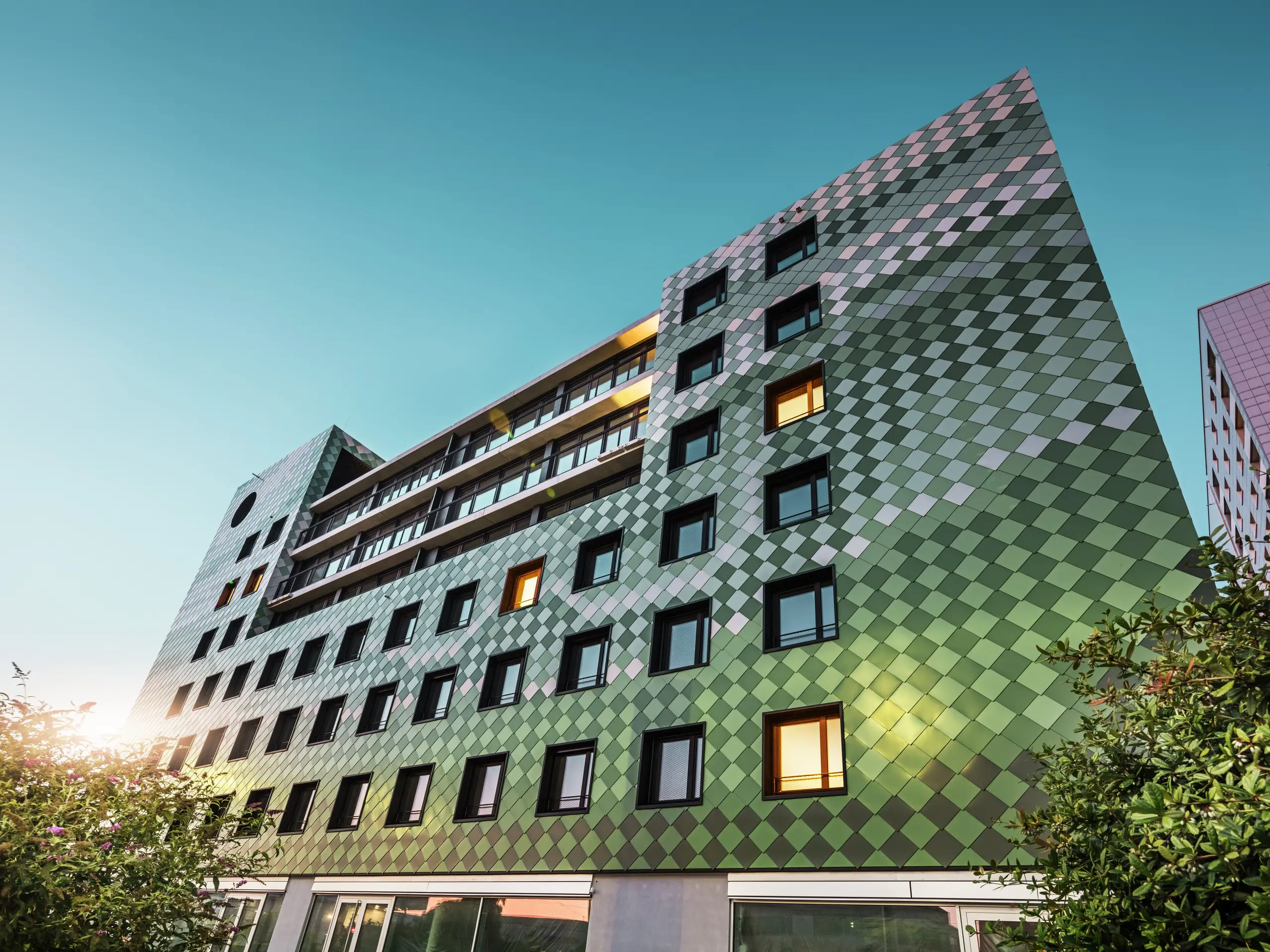 Bâtiment résidentiel et commercial moderne "Pick-Nick" à Dresde avec une façade PREFA frappante en losanges d'aluminium dans des couleurs spéciales vert olive, vert gris, vert réséda, vert opale, ivoire clair, blanc perle et bronze clair. Le traitement créatif de la façade rappelle une rizière et crée un jeu de couleurs vivant. L'architecture claire, les cadres de fenêtres noirs et la durabilité des matériaux PREFA façonnent le bâtiment. Le soleil bas se reflète sur la façade et les surfaces des fenêtres.