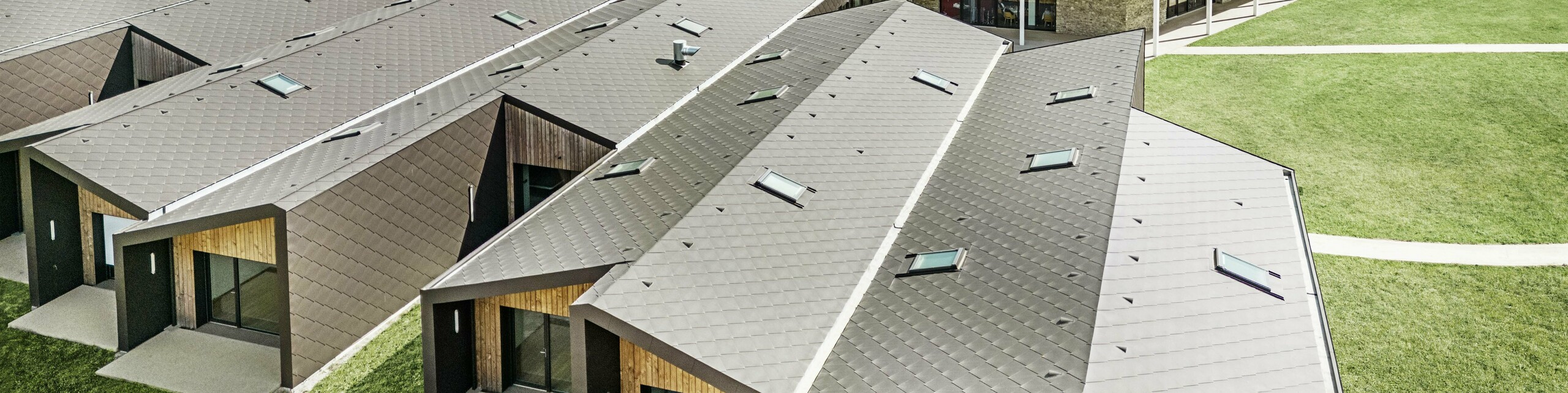 Slika prikazuje zračnu snimku modernog stambenog kompleksa, obloženog PREFA krovnim i zidnim rombovima 44 × 44 u orah smeđoj boji. Krovni pejzaž karakteriziraju geometrijski oblici, koji su kombinirani s velikim staklenim površinama i drvenim elementima. U pozadini se nalazi stara crkva, koja stvara skladan kontrast s modernom arhitekturom. Zeleni travnjak i okolna priroda dodatno naglašavaju otvoren i održiv dizajn cijelog kompleksa.