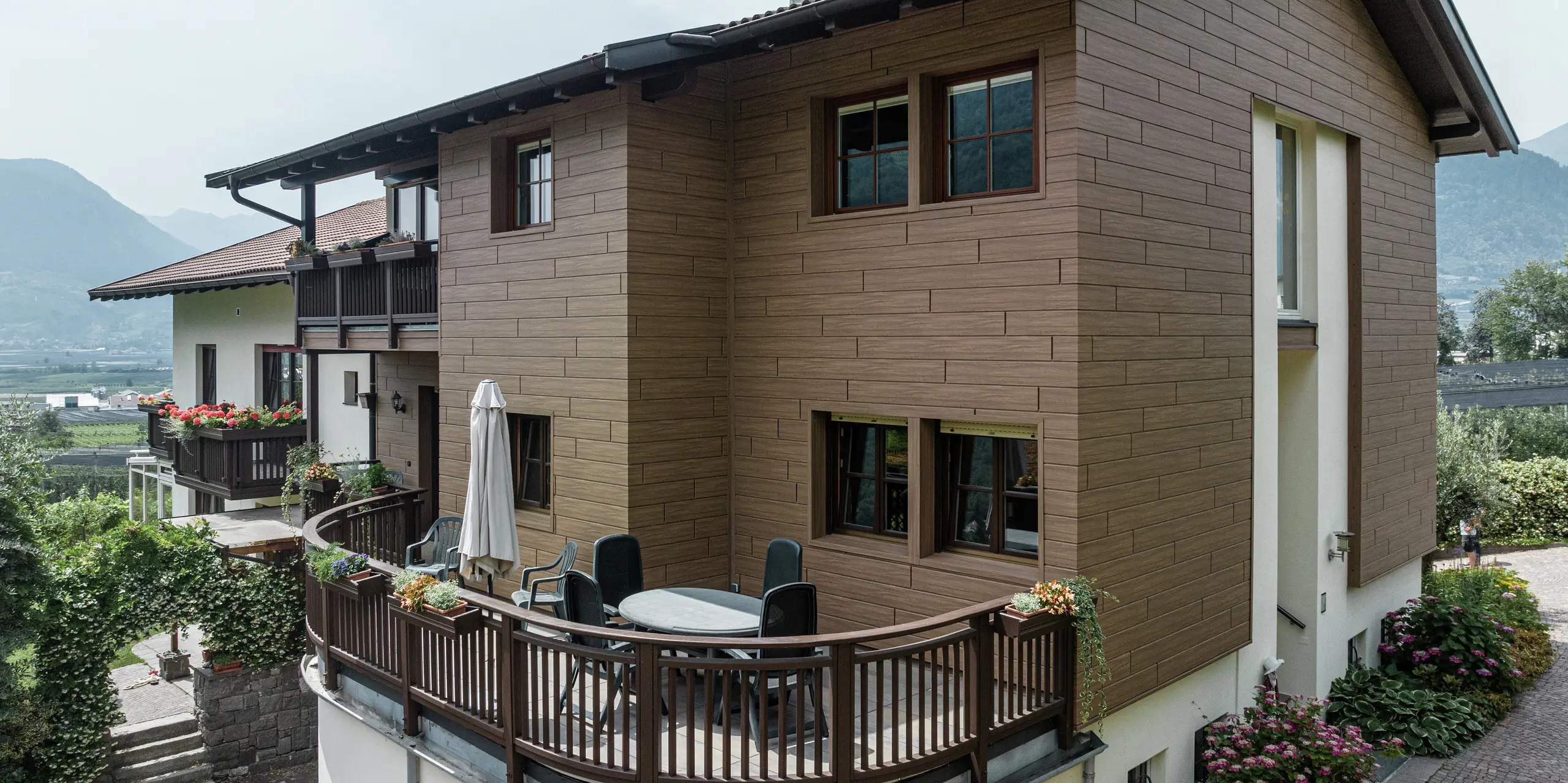 Appartements de vacances rénovés Prechtlhof à Merano, Tyrol du Sud, avec sIDING PREFA en noyer foncé. La façade en aluminium sans entretien à l'aspect bois allie esthétique moderne et charme traditionnel et s'intègre harmonieusement dans l'environnement alpin. Le balcon circulaire au premier plan vous invite à vous attarder dans le magnifique paysage du Tyrol du Sud.