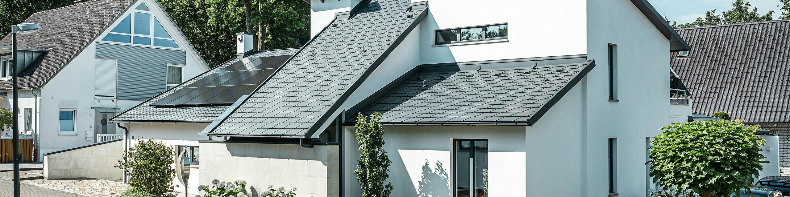 Modernes Einfamilienhaus mit weißer Putzfassade und dunklem PREFA Schrägdach aus Aluminium. Auf dem Dach sind integrierte Photovoltaikmodule und eine Aluminium-Dachentwässerung in Anthrazit zu sehen. Das Gebäude liegt in einer gepflegten Wohnsiedlung mit bepflanzten Vorgärten, gepflasterter Zufahrt und umliegenden Einfamilienhäusern.