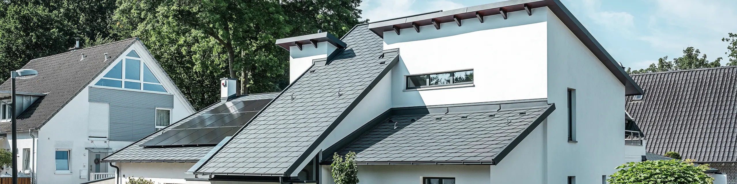 Moderne eengezinswoning met witte gepleisterde gevel en donker PREFA-hellend dak van aluminium. Op het dak zijn geïntegreerde fotovoltaïsche modules en aluminium dakafvoersystemen in antraciet te zien. Het gebouw ligt in een verzorgde woonwijk met beplante voortuinen, een geplaveide oprit en omliggende eengezinswoningen.