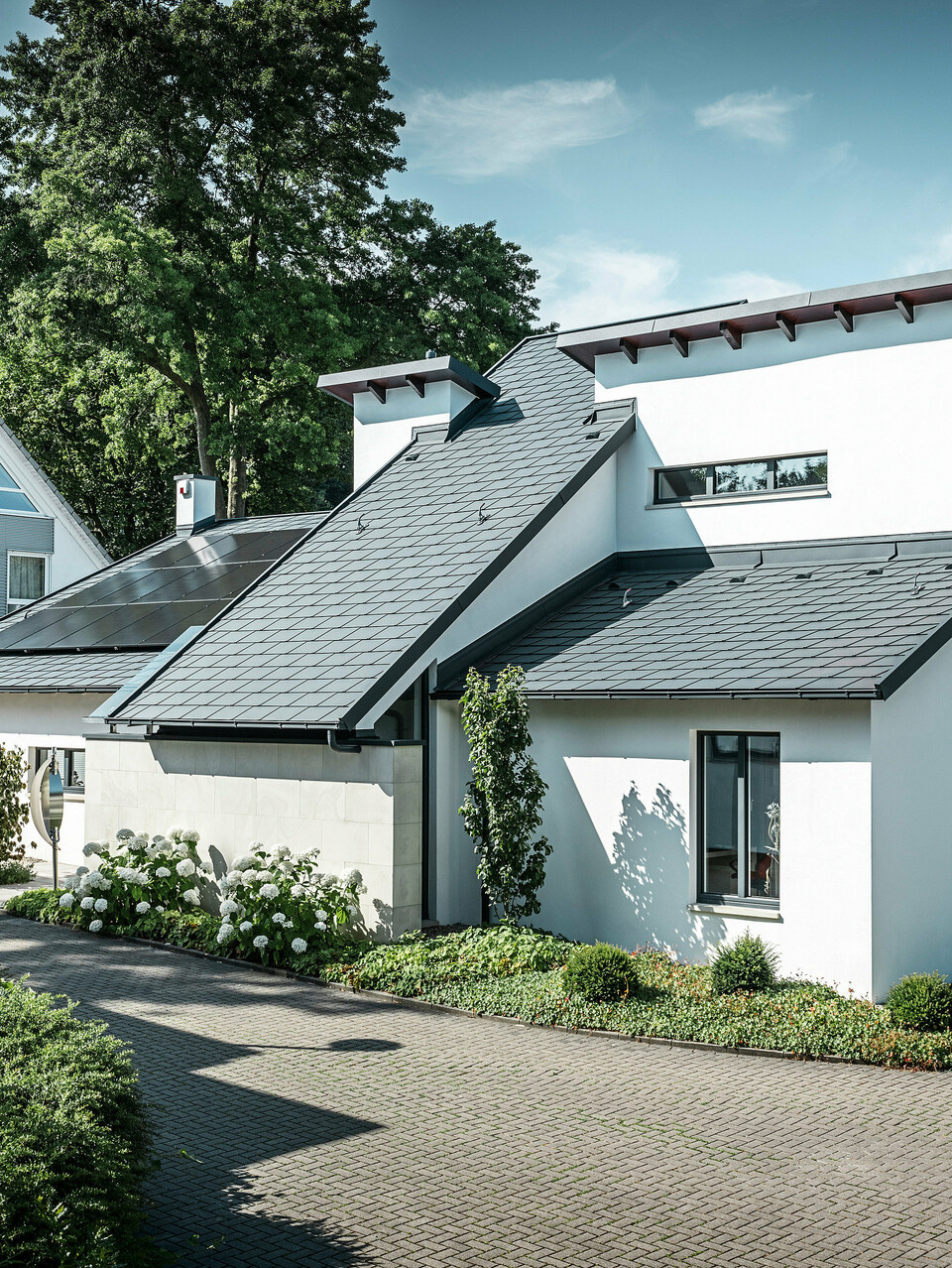 Modernes Einfamilienhaus mit weißer Putzfassade und dunklem PREFA Schrägdach aus Aluminium. Auf dem Dach sind integrierte Photovoltaikmodule und eine Aluminium-Dachentwässerung in Anthrazit zu sehen. Das Gebäude liegt in einer gepflegten Wohnsiedlung mit bepflanzten Vorgärten, gepflasterter Zufahrt und umliegenden Einfamilienhäusern.