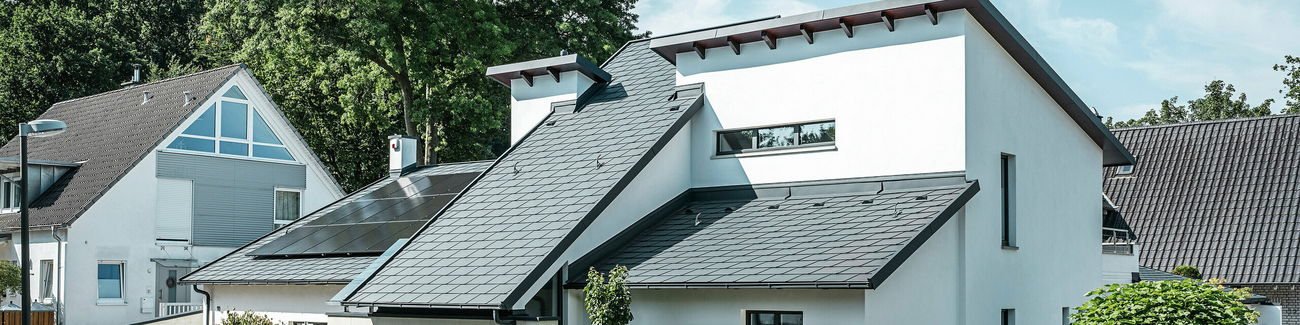 Maison individuelle moderne avec façade en crépi blanc et toit en pente PREFA en aluminium foncé. La toiture est équipée de modules photovoltaïques intégrés et de gouttières en aluminium anthracite. Le bâtiment est situé dans un lotissement bien entretenu avec des jardins fleuris, une allée pavée et des maisons individuelles environnantes.