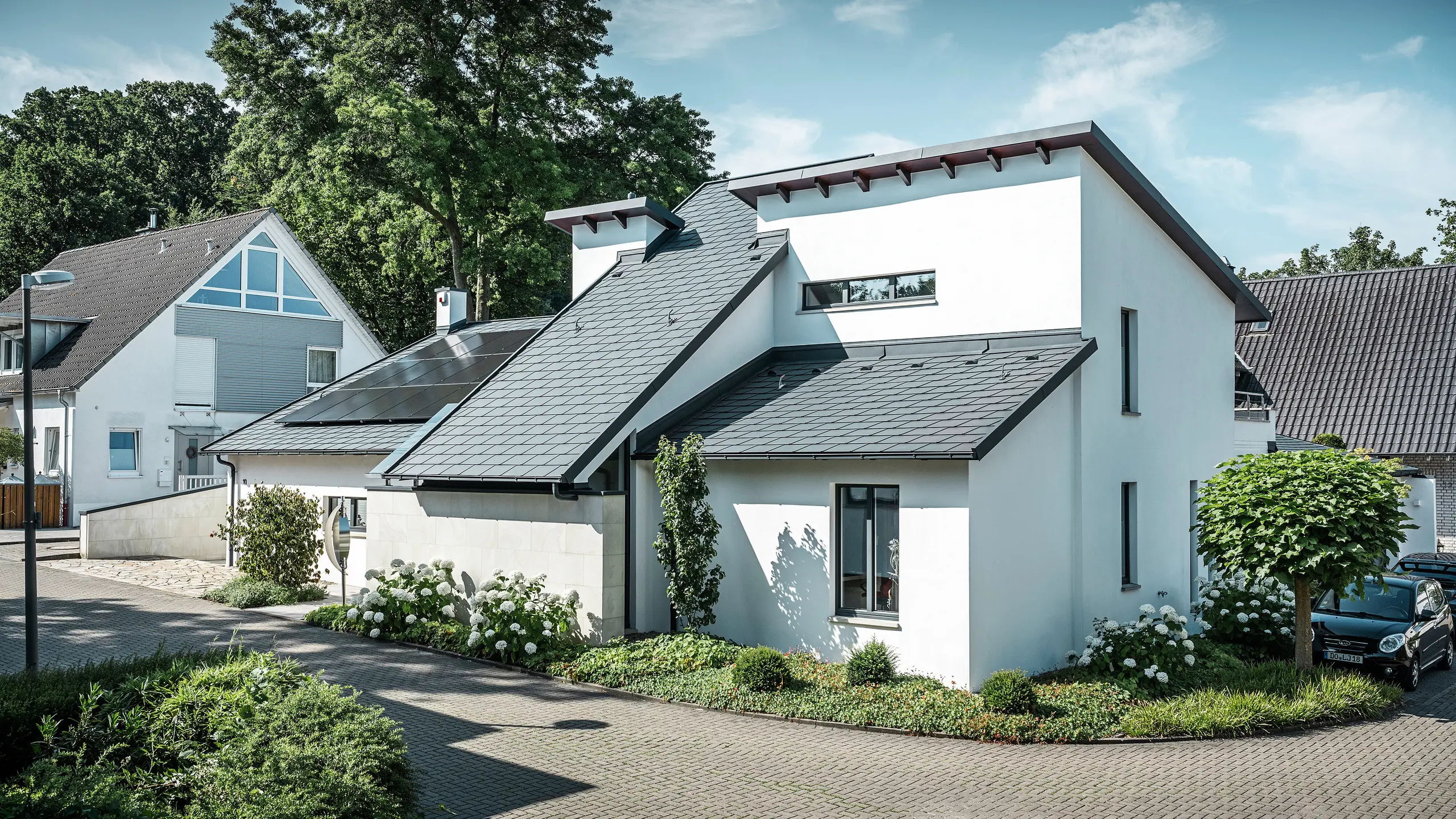 Moderne eengezinswoning met witte gepleisterde gevel en donker PREFA-hellend dak van aluminium. Op het dak zijn geïntegreerde fotovoltaïsche modules en aluminium dakafvoersystemen in antraciet te zien. Het gebouw ligt in een verzorgde woonwijk met beplante voortuinen, een geplaveide oprit en omliggende eengezinswoningen.