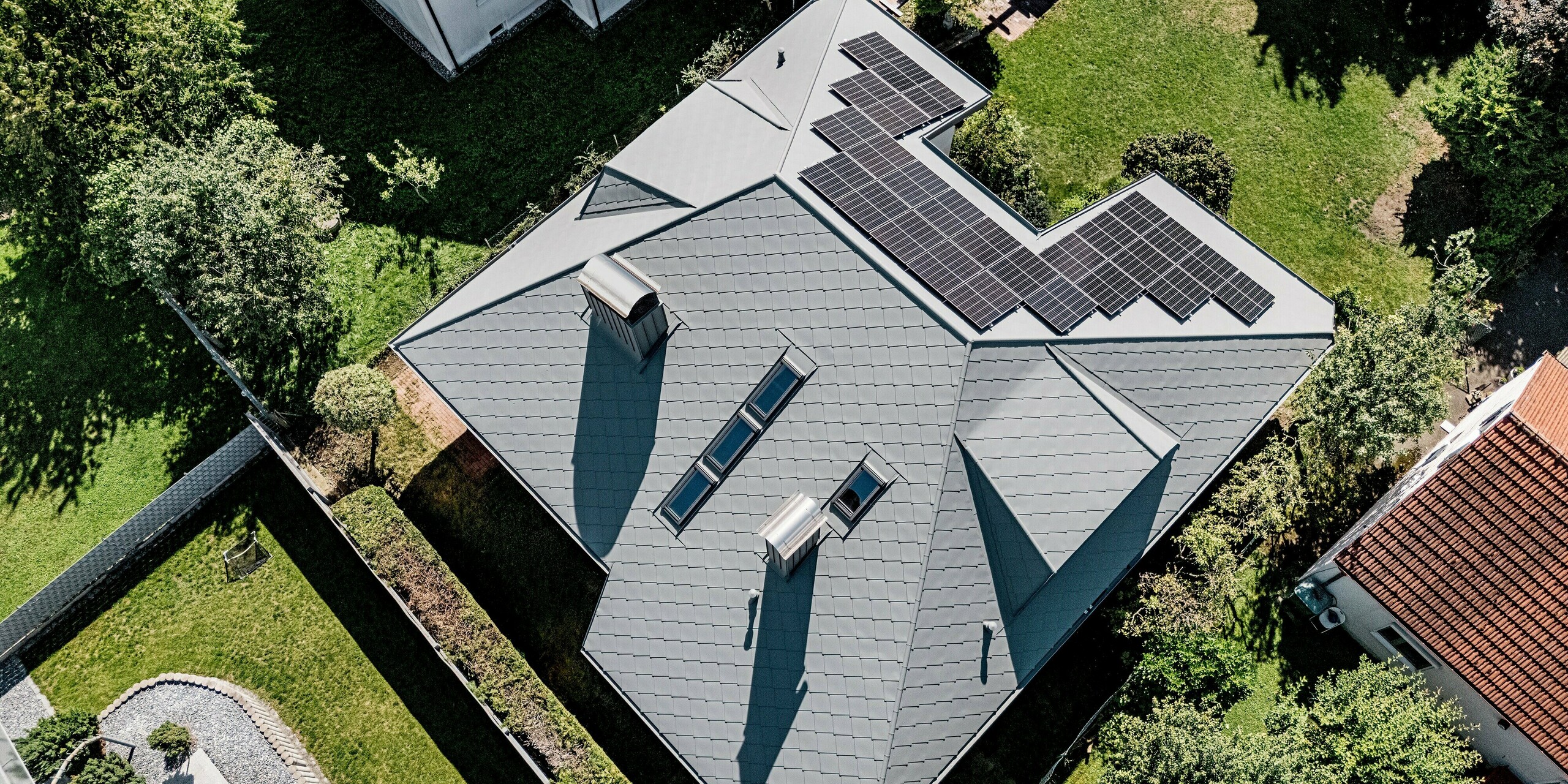 Das Bild zeigt eine beeindruckende Vogelperspektive auf ein Walmdach, das vollständig mit PREFA Dachrauten 44 × 44 in P.10 Hellgrau eingedeckt ist. Die markante Rautenstruktur und die klaren Linien der Trapezgauben, Dachfenster und Kaminverkleidungen verleihen dem Dach eine moderne, präzise Optik. Die großflächig integrierte Photovoltaikanlage wirkt kraftvoll und effizient. Eingebettet in einen grünen Garten zeigt das langlebige, witterungsbeständige PREFA Aluminiumdach seine Stärke besonders deutlich – stabil, formschön und perfekt verarbeitet.