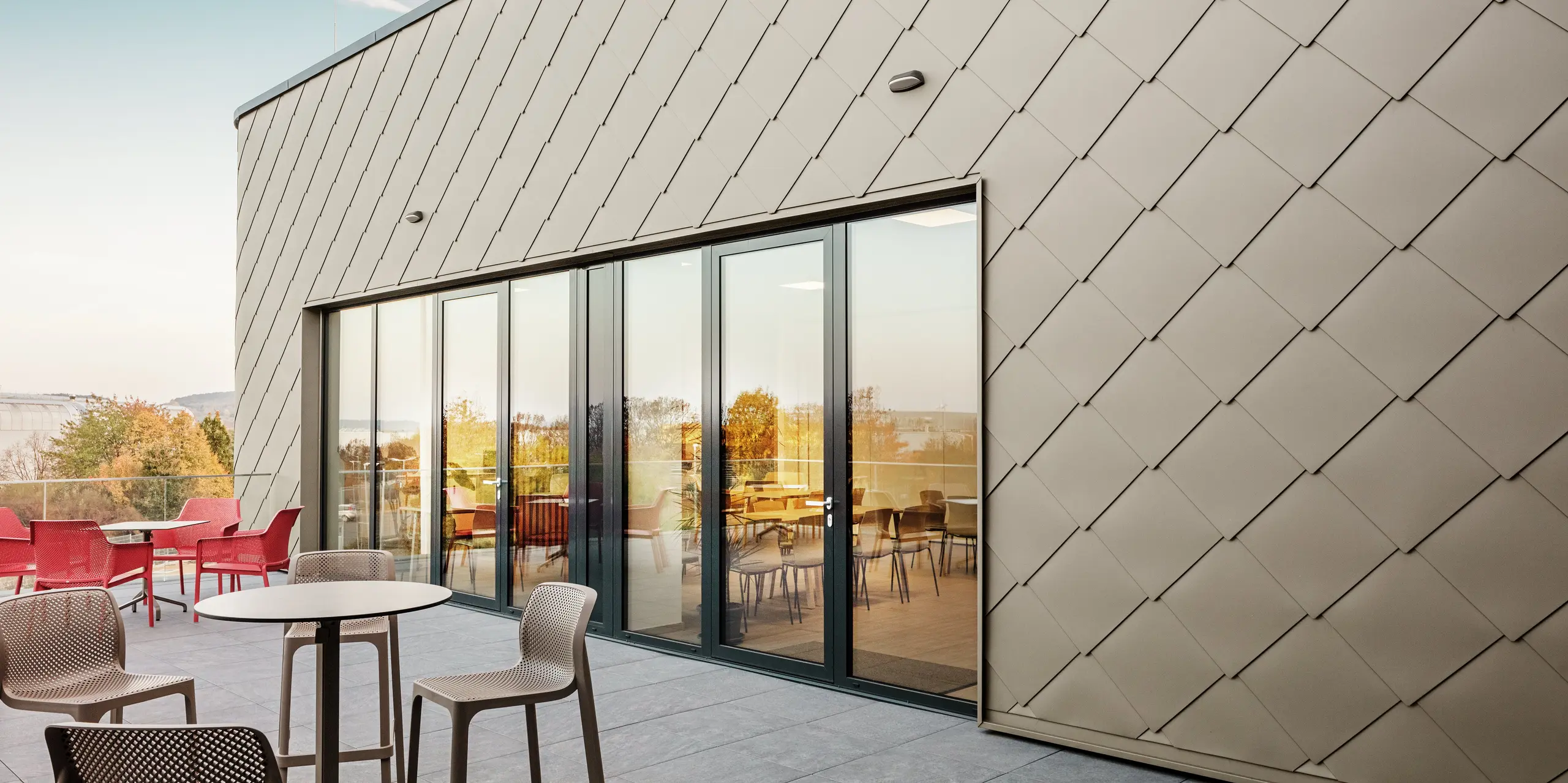 La façade du nouveau site PREFA à Törökbálint impressionne par les élégants losanges de façade PREFA 44 × 44 en P.10 bronze prefa. La façade en aluminium posée avec précision crée une apparence moderne et dynamique tout en offrant une protection maximale contre les éléments. La généreuse façade vitrée relie les espaces intérieurs et extérieurs et crée une atmosphère lumineuse et accueillante sur la terrasse spacieuse.