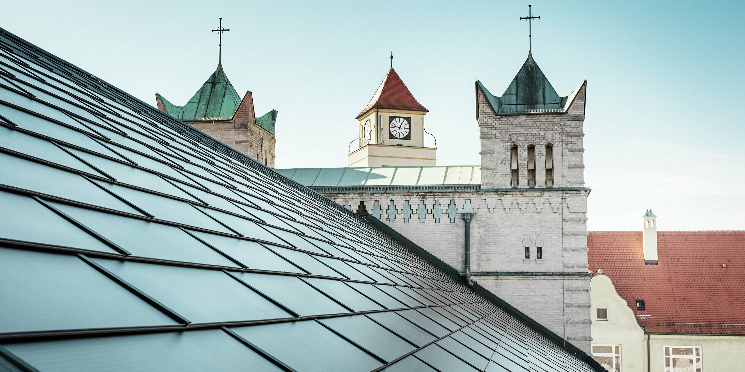 Le toit remarquable de l'église Saint-Charles-Borromée de Nuremberg a été recouvert de tuiles solaires PREFA noires dans le cadre d'une rénovation complète. La vue détaillée du toit à deux versants et des imposants clochers en arrière-plan témoigne de l'intégration réussie de la technologie solaire moderne dans l'architecture historique de l'église. Les tuiles solaires fournissent une énergie durable et s'intègrent harmonieusement à l'architecture traditionnelle. C'est un nouvel exemple de l'alliance réussie entre préservation du patrimoine historique et production d'énergie innovante. Un ciel bleu éclatant se dessine en arrière-plan.