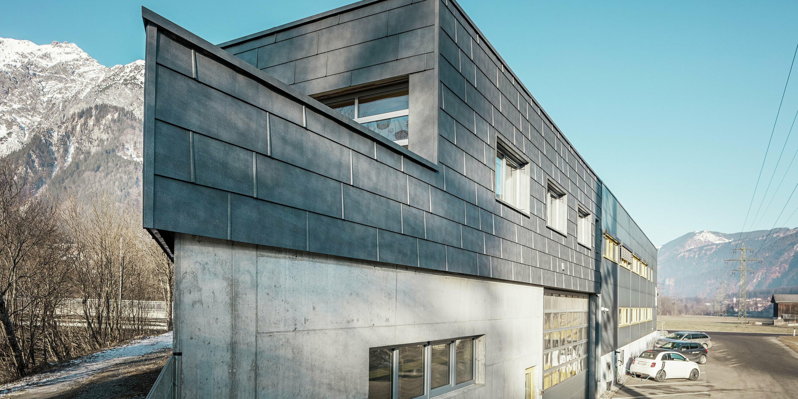 Façade du bâtiment de l'entreprise de couverture RÜMA, dans le Vorarlberg. Sa façade remarquable, composée de panneaux en aluminium PREFA FX.12 et d'une ossature en béton apparente, s'intègre parfaitement dans un paysage alpin, avec montagnes et ciel bleu. Une voiture électrique en charge est visible sur le parking devant le bâtiment, soulignant ainsi la durabilité de l'entreprise.