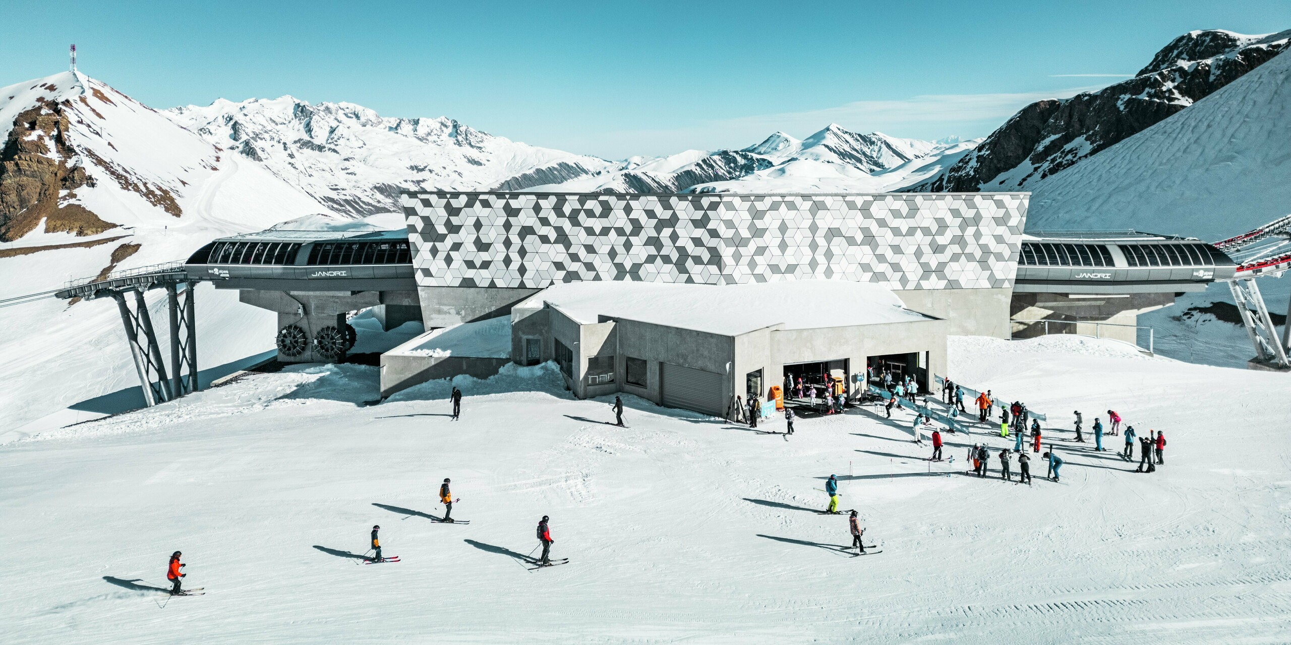La station de téléphérique futuriste des Deux Alpes, nichée au cœur d'un paysage montagneux époustouflant, arbore une façade remarquable en panneaux composites en aluminium PREFABOND. Son motif géométrique saisissant en losanges blancs et gris s'intègre parfaitement à l'environnement enneigé. Les matériaux durables et résistants aux intempéries offrent une solution esthétique et fonctionnelle pour les conditions extrêmes de la haute montagne. De nombreux skieurs et snowboarders se préparent pour la descente devant la station intermédiaire.