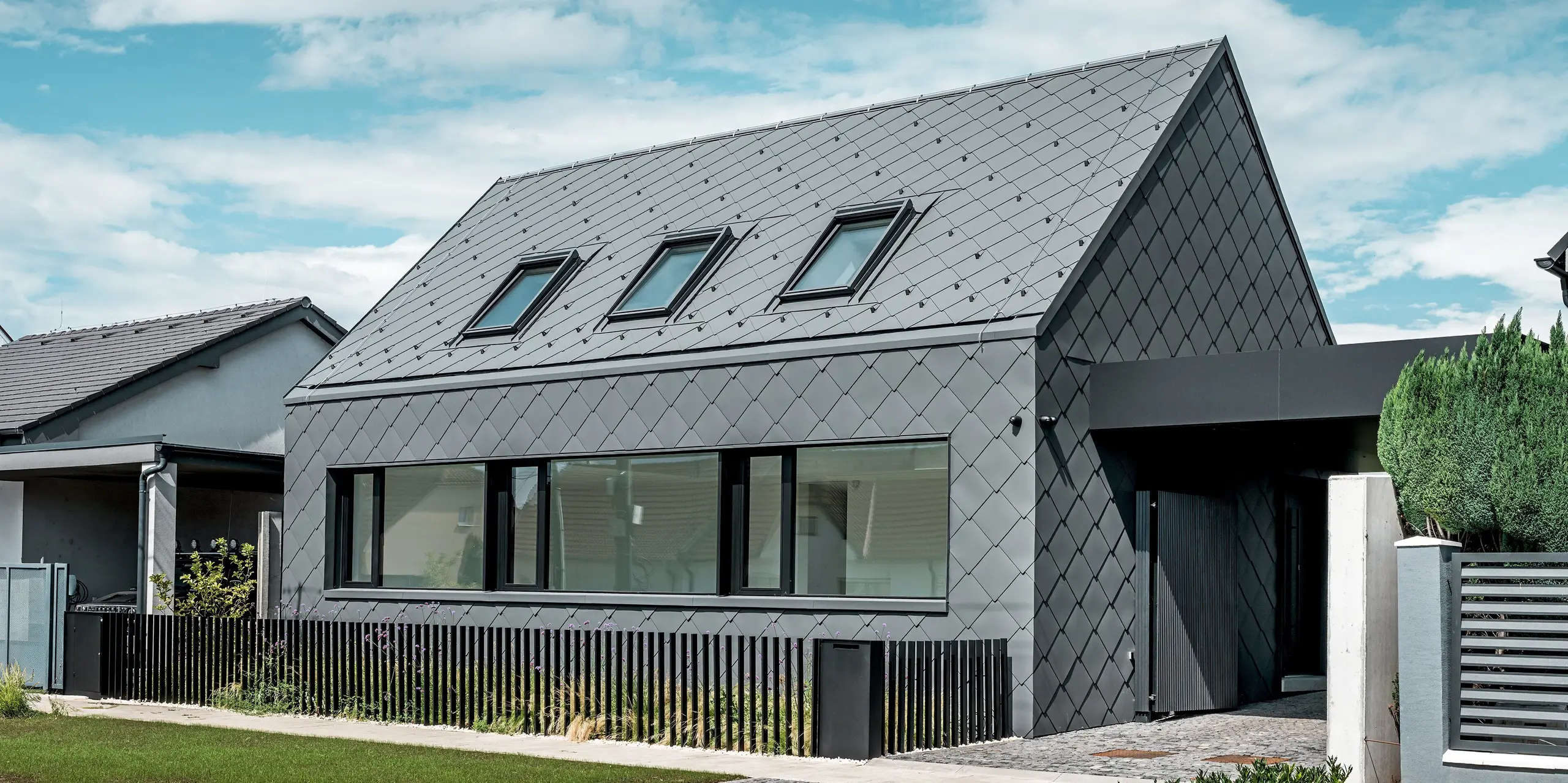 Maison individuelle moderne à Trnava-Modranka avec bardage aluminium : l’enveloppe de cette structure minimaliste est entièrement revêtue de losanges 44x44 PREFA P.10 gris sombre, utilisées pour la toiture et les façades. Le motif losange, saisissant, se déploie harmonieusement sur le toit et la façade, conférant à la maison une allure monolithique et affirmée. De larges fenêtres soulignent l’architecture, tandis que l’aluminium PREFA, résistant aux intempéries, assure une protection durable et une élégante finition mate. Le ciel lumineux accentue le contraste avec l’enveloppe sombre du bâtiment et met en valeur son design moderne.