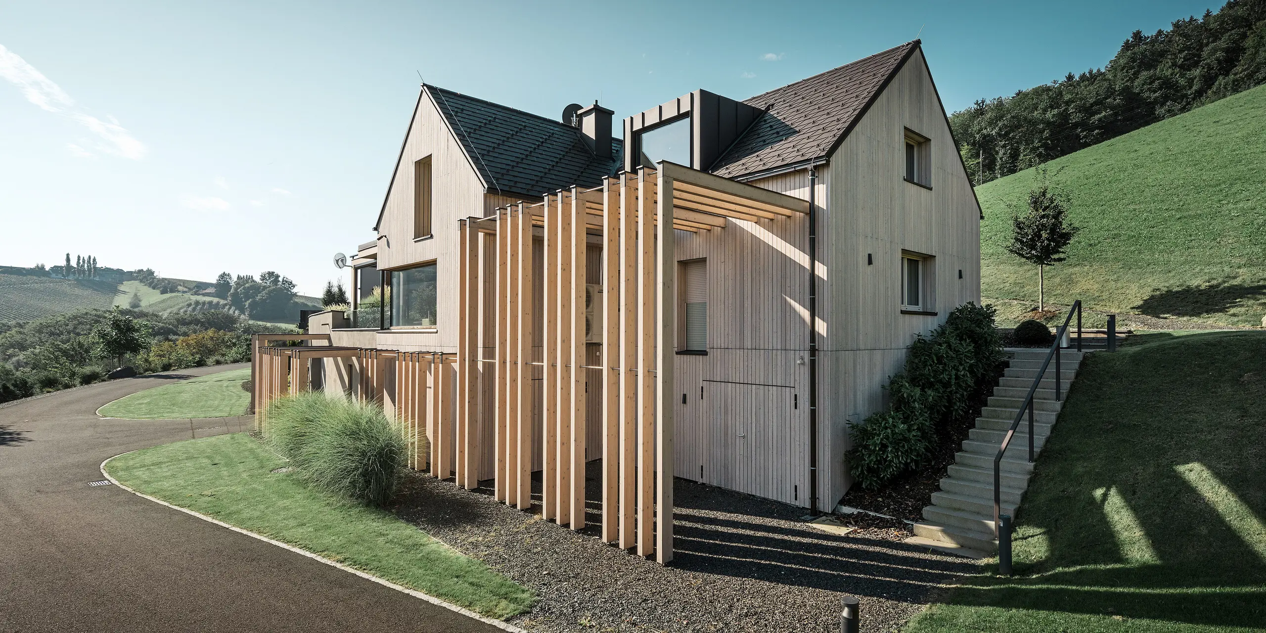 Grote eengezinswoning met zadeldak en dakkapel op een helling. Het dak is bedekt met bruine PREFA aluminium dakspanen en de gevel is bekleed met verticale houten planken.