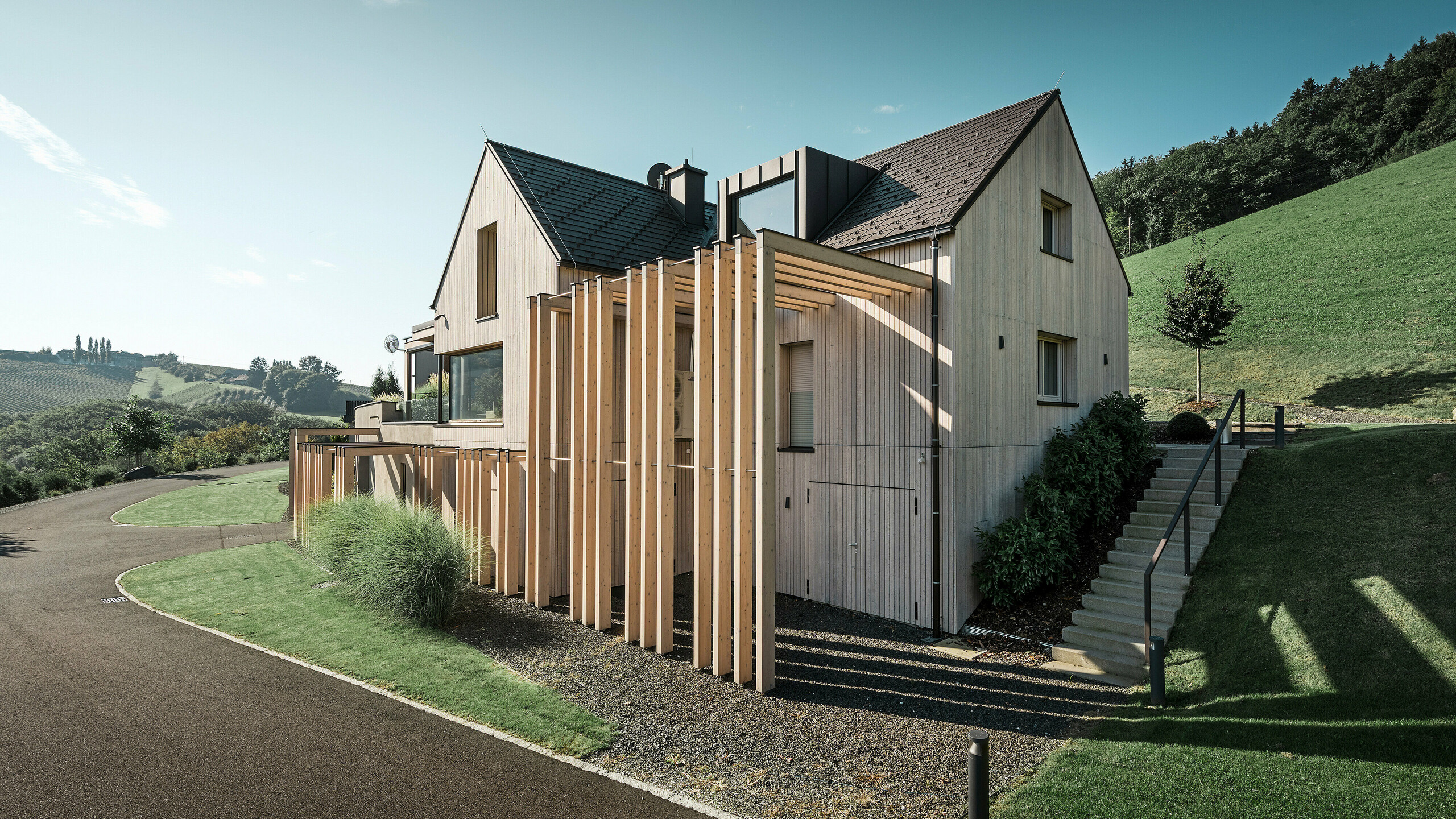 Grote eengezinswoning met zadeldak en dakkapel op een helling. Het dak is bedekt met bruine PREFA aluminium dakspanen en de gevel is bekleed met verticale houten planken.