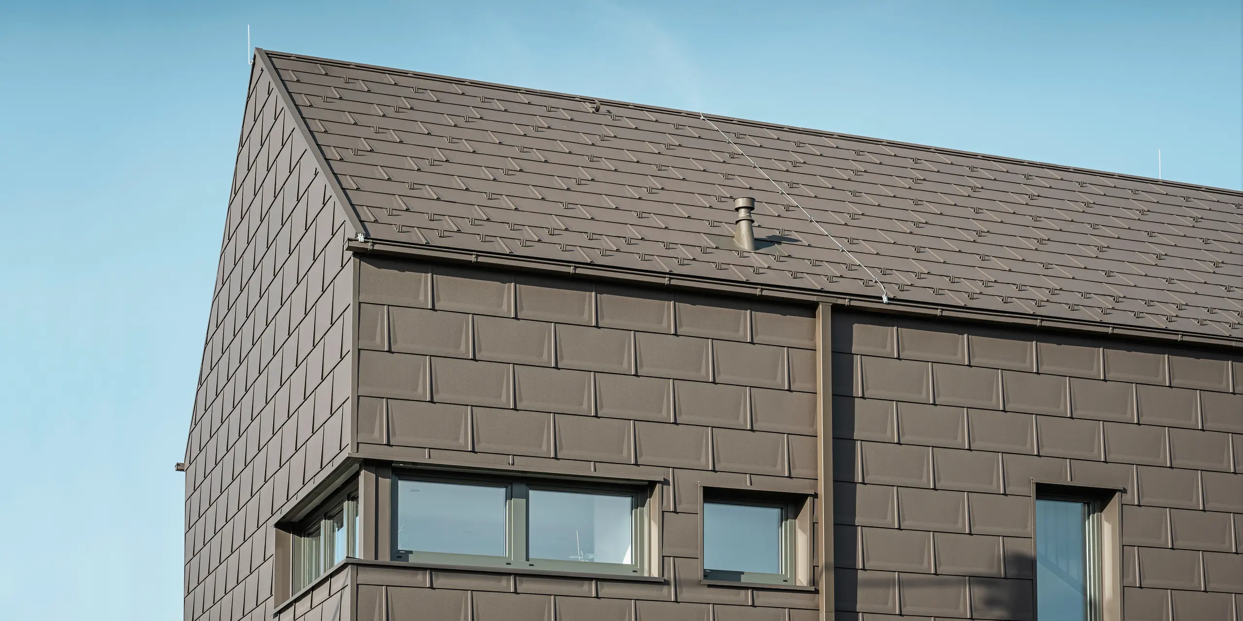 Detailaufnahme eines Einfamilienhauses in Neukirchen, Österreich, mit einer Dach- und Fassadenverkleidung aus PREFA R.16 Dachplatten in P.10 braun. Die präzise Überlappung der Platten sorgt für ein einheitliches und ansprechendes Erscheinungsbild, das moderne Ästhetik mit Funktionalität verbindet. Die integrierte PREFA Dachentwässerung in Form einer Kastenrinne und eines Vierkantrohrs in P.10 Braun zeigt das Engagement von PREFA für hochwertige und optisch ansprechende Bauelemente.