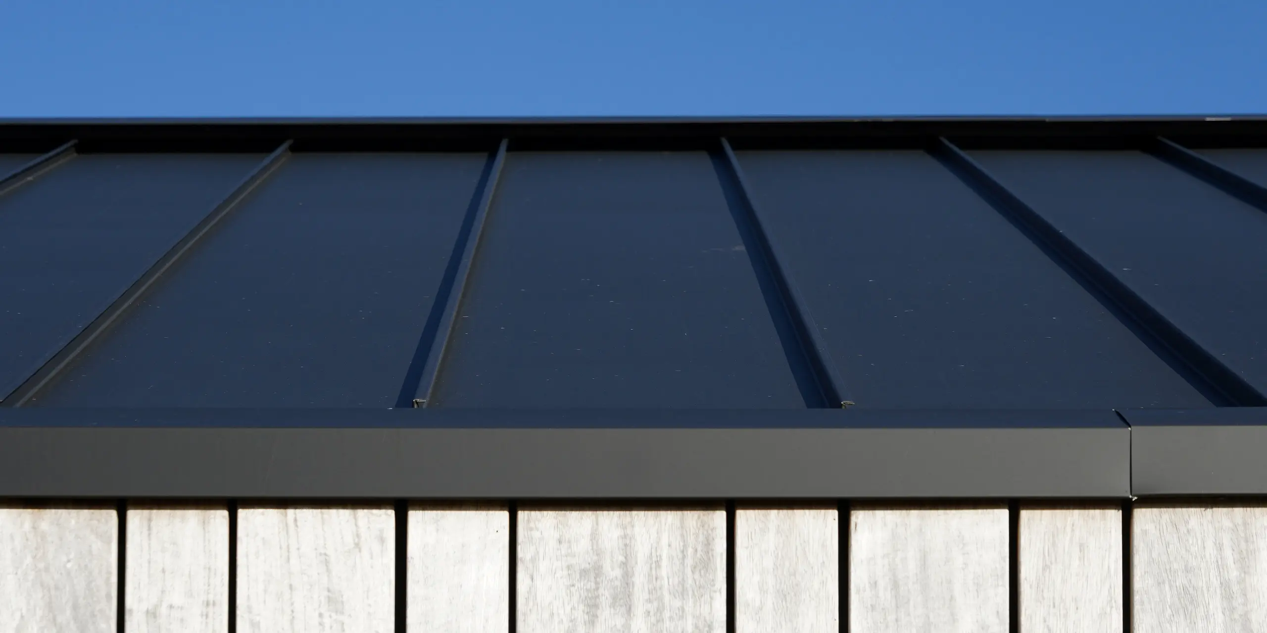 Vue détaillée d'une maison familiale moderne à Uithoorn, aux Pays-Bas, avec toiture à joints debout PREFALZ en gris noir. L'image montre le traitement précis de l'aluminium de haute qualité de PREFA, combinée à des éléments en bois naturel sur la façade. Les lignes claires du toit robuste en aluminium soulignent la construction durable et résistante aux intempéries.