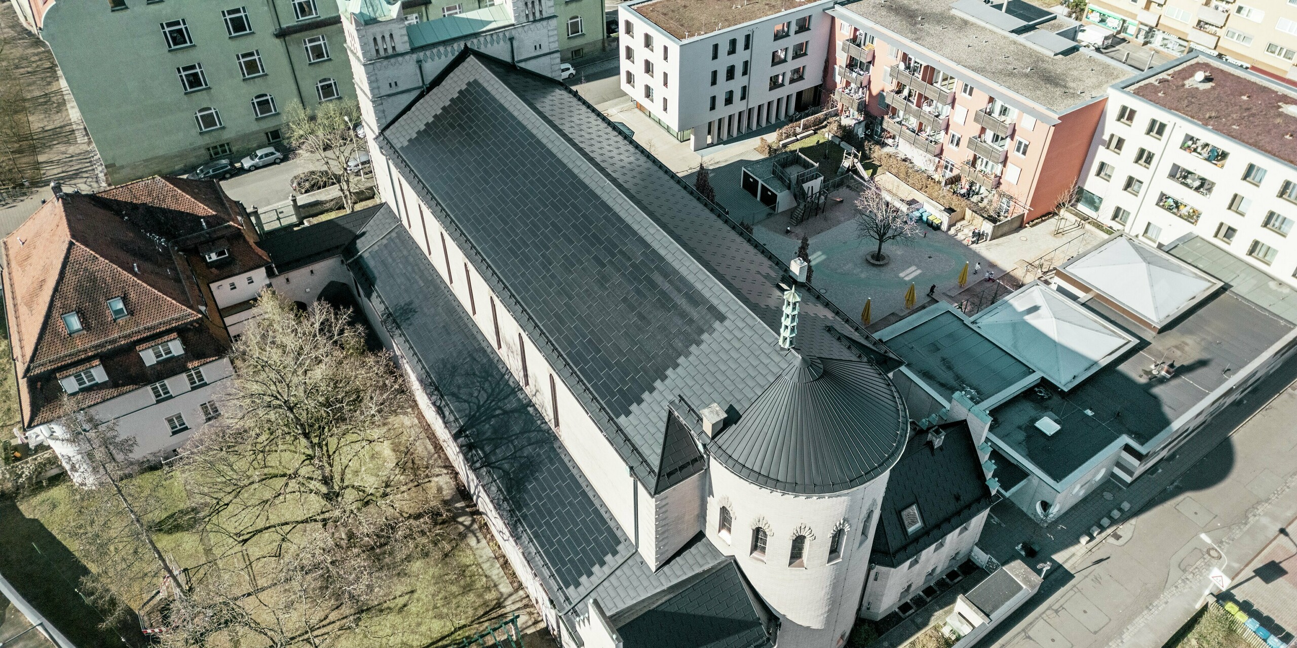 Vue aérienne de l'arrière de l'église Saint-Charles-Borromée de Nuremberg. Des tuiles solaires PREFA noires recouvrent le remarquable toit à pignon arrondi qui surplombe le corps principal de l'église, ainsi que celui du bâtiment adjacent. Les panneaux de toiture en aluminium avec modules photovoltaïques intégrés s'intègrent parfaitement à l'aspect historique de l'église. La vue sur l'environnement moderne, avec ses immeubles d'habitation adjacents et sa cour intérieure verdoyante, illustre l'alliance réussie entre préservation du patrimoine, architecture moderne et production d'énergie durable. Une solution idéale pour la préservation du patrimoine en milieu urbain.