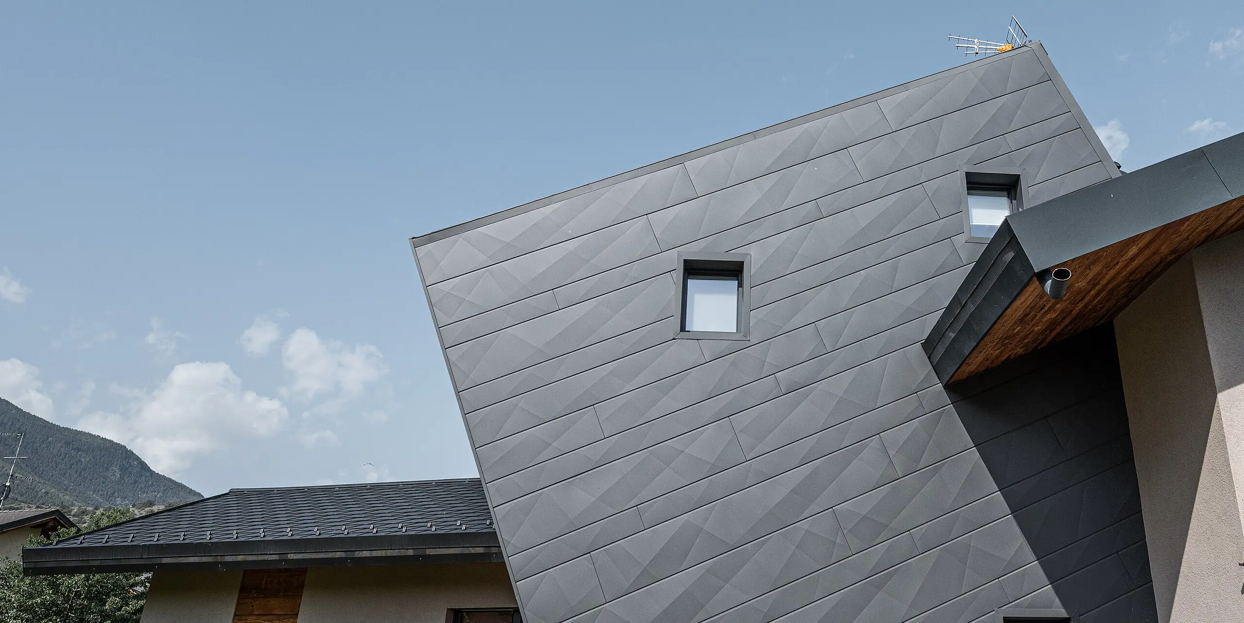Die Villa Tia in Gressan, Italien, zeigt eine moderne Fassadengestaltung mit PREFA Siding.X in P.10 Dunkelgrau. Die dynamische Anordnung der Paneele erzeugt ein dreidimensionales Erscheinungsbild, das durch den Kontrast zu den natürlichen Holzelementen und der umgebenden Landschaft hervorgehoben wird. Diese innovative Sonderlösung der schrägen Siding.X Fassadenpaneele steht für ein zeitgemäßes und langlebiges Design.