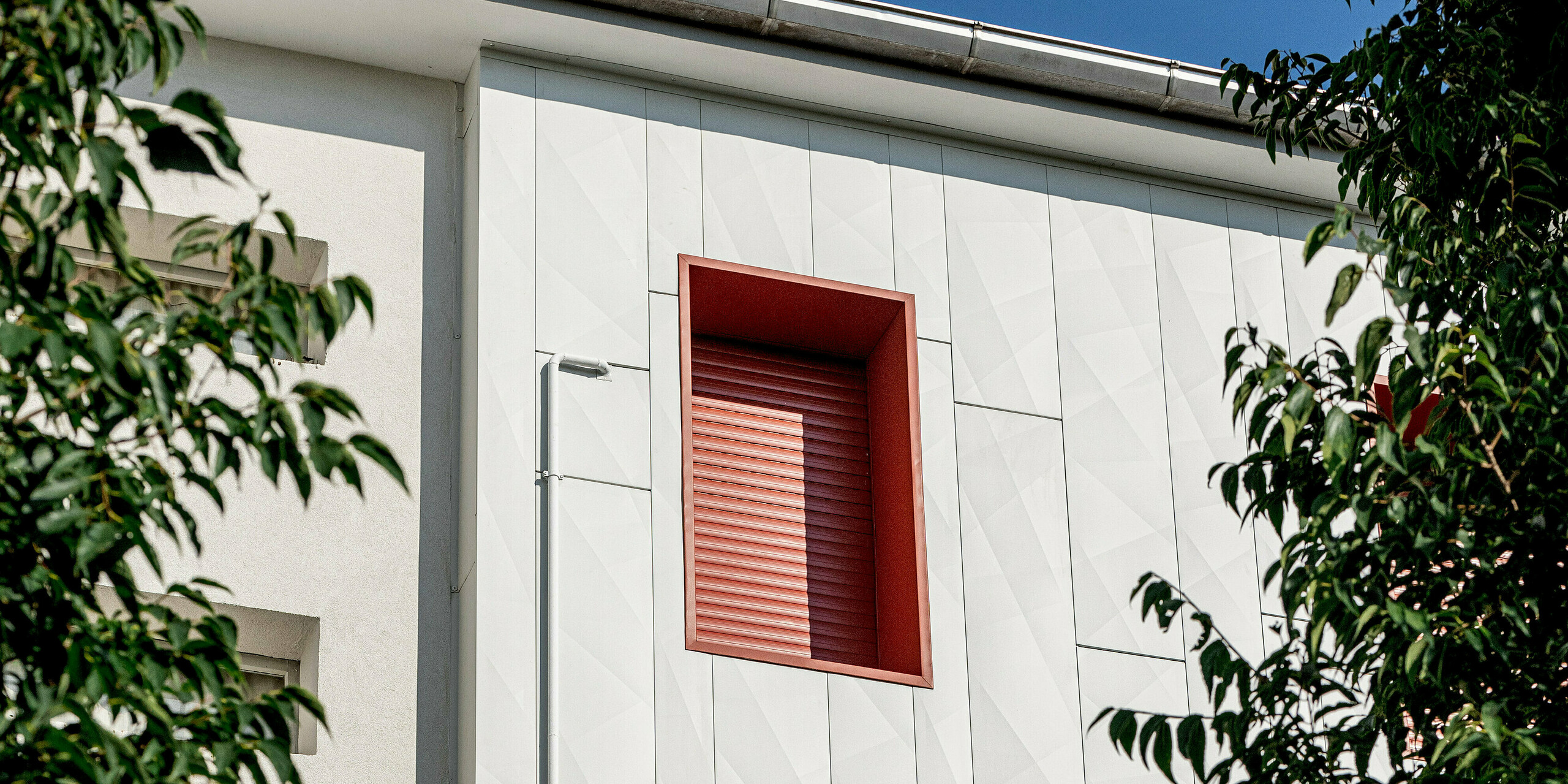 Moderne Wohnanlage Alba B nach der Sanierung mit PREFA Siding.X Aluminiumfassade: Die vertikal verlegten Paneele erzeugen spannende Licht- und Schattenspiele auf der hellen Oberfläche. Markante rote Fensterrahmen setzen starke Kontraste und lenken den Blick gezielt auf die Fassadenstruktur. Umgeben von grünen Baumkronen wirkt das Gebäude lebendig und hochwertig. Die PREFA Aluminiumfassade überzeugt durch Langlebigkeit, Widerstandsfähigkeit und Wartungsarmut und bietet zugleich eine zeitgemäße, architektonisch prägnante Optik.