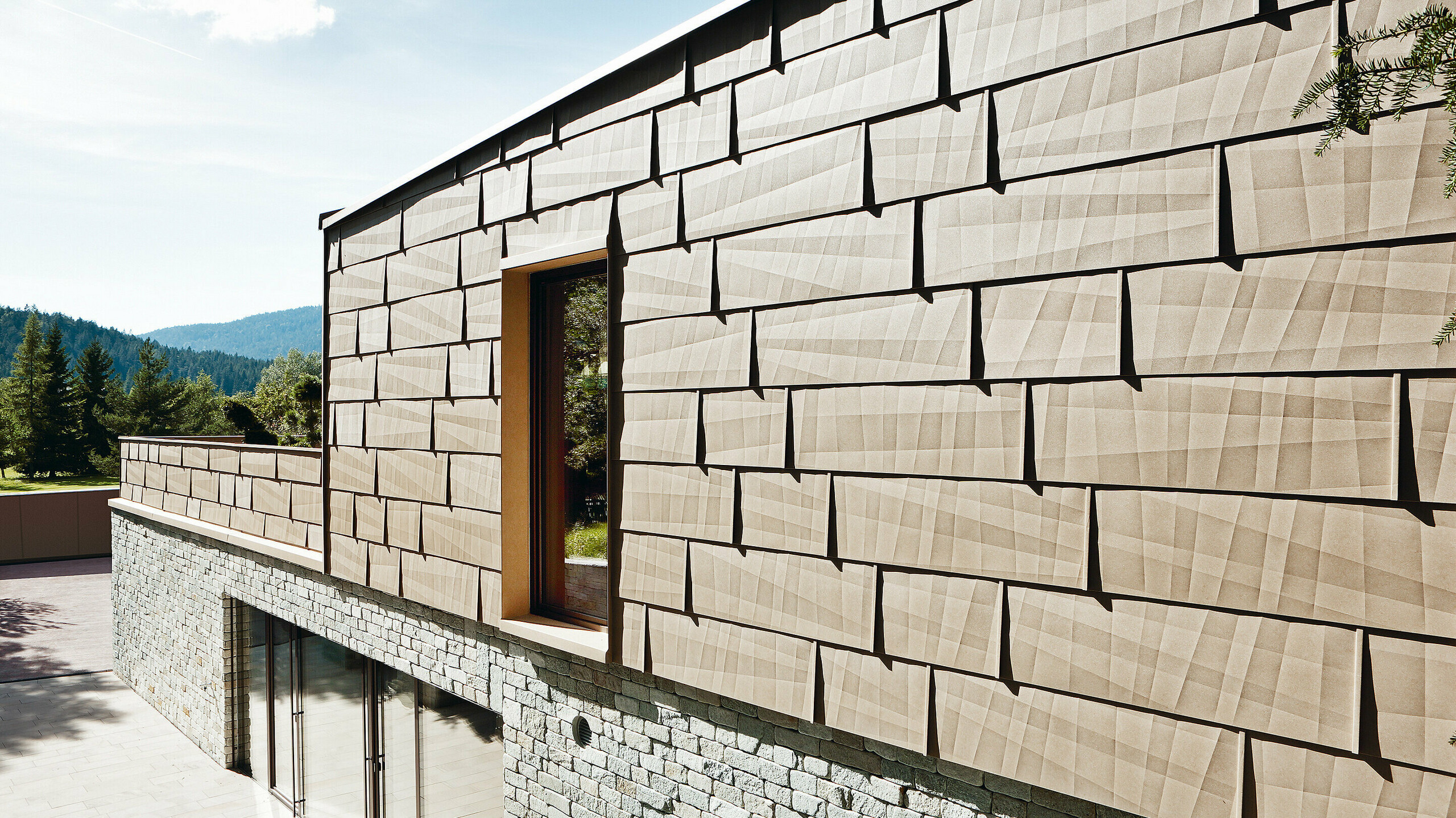 An dieser Außenfassade wurde das PREFA Fassadenpaneel FX.12 in P.10 Sandbraun mit einer Steinfassade im Untergeschoß kombiniert.