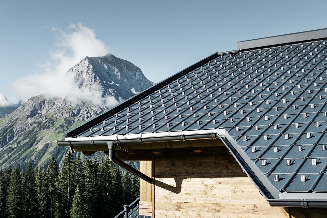 Tuile PREFA employée en région montagneuse - cabane recouverte avec des tuiles anthracite à Lech am Arlberg avec montagnes en arrière-plan ; gouttière PREFA anthracite ; façade en bois