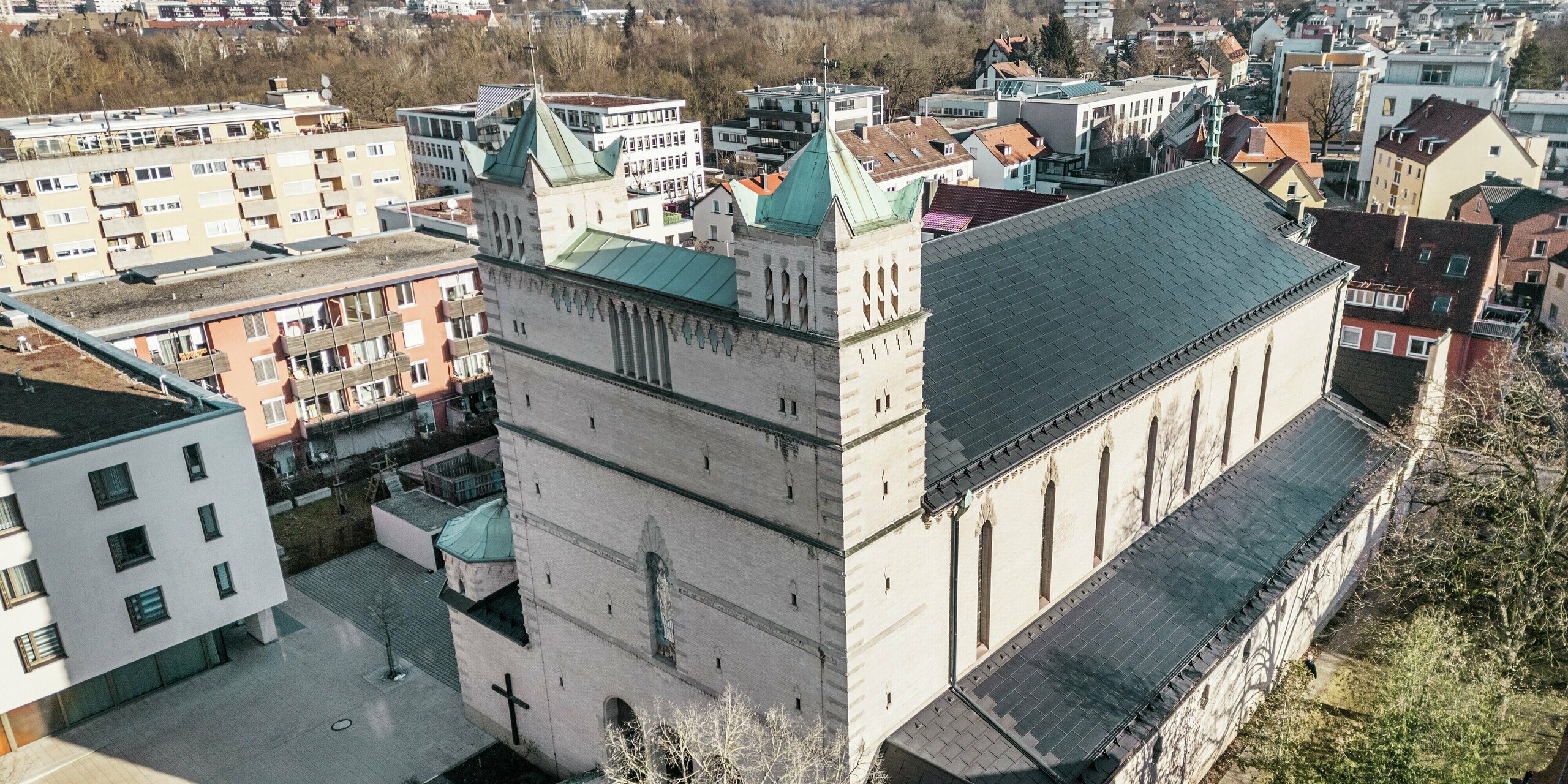 Façade de l'église Saint-Charles-Borromée de Nuremberg : l'image montre l'impressionnant toit de cette église classée, recouvert de tuiles solaires PREFA noires. Ces modules s'intègrent harmonieusement à l'architecture historique de l'église tout en représentant une solution moderne pour une production d'énergie durable. En arrière-plan, on aperçoit des bâtiments modernes et les collines verdoyantes de Nuremberg, soulignant la transition réussie entre l'architecture religieuse traditionnelle et l'approvisionnement énergétique contemporain. La toiture en aluminium de l'église est complétée par l'élégant R.16 PREFA.