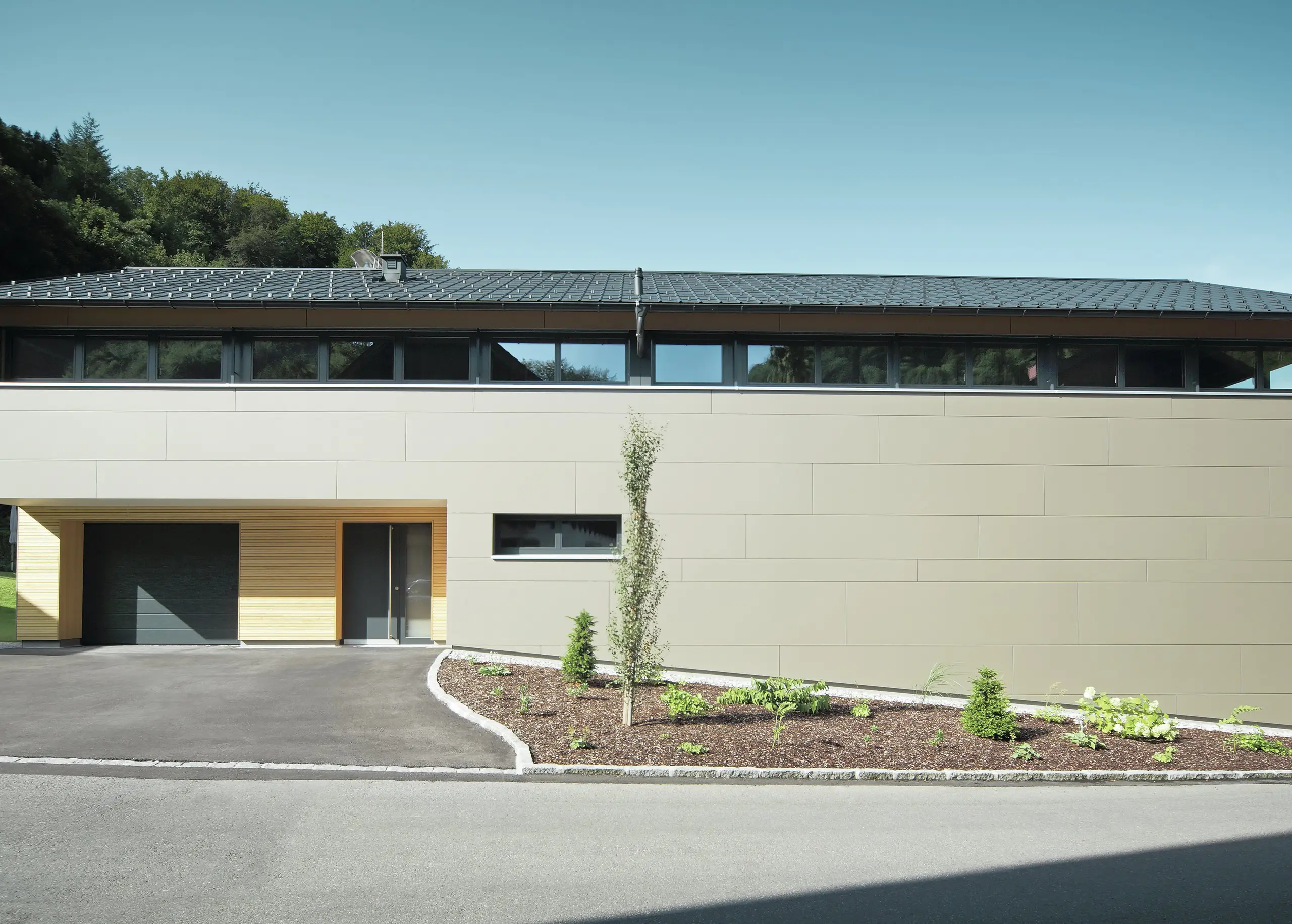 Das Einfamilienhaus in Hohenems präsentiert sich in einer modernen und stilvollen Fassade aus PREFA Aluminium Verbundplatten in Bronzemetallic, mit einem darüberliegenden Dach aus PREFA Dachplatten in P.10 Anthrazit. Die klare Linienführung und das durchdachte Farbkonzept des Designs integrieren das Gebäude nahtlos in die natürliche Umgebung. Die Gestaltung zeigt die Kombination aus Funktionalität und Ästhetik, die PREFA Produkte für zeitgemäße Wohnarchitektur bieten.